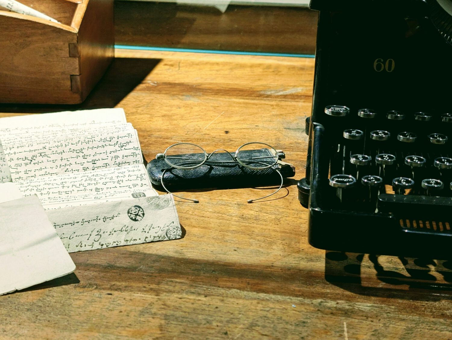 Vintage black typewriter sits on a wooden desk next to wire-rimmed glasses resting on a dark leather case. Handwritten papers with cursive script and official seals are scattered nearby, creating an atmospheric writer's workspace scene.