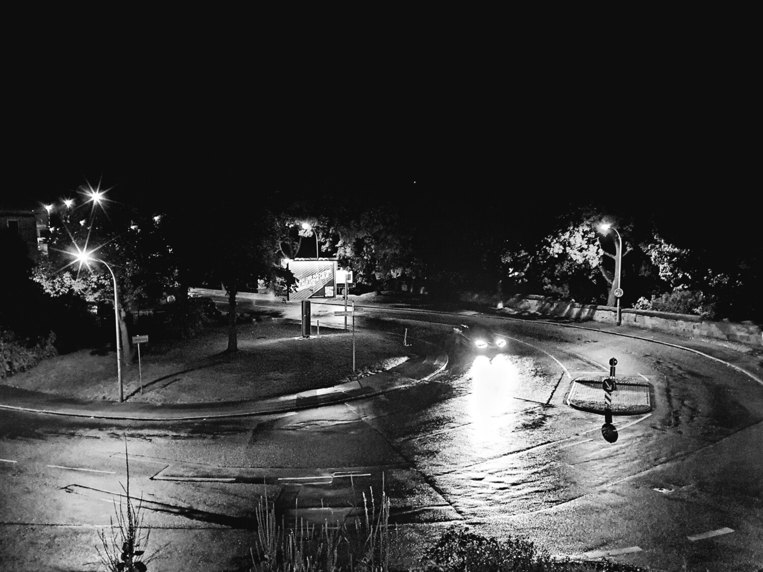 Night view of a curved road intersection, illuminated by street lamps. A car's headlights shine on the wet pavement, creating a dramatic contrast in the black and white image. Trees and road signs are visible in the shadowy surroundings.