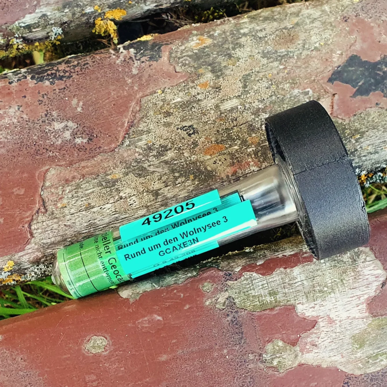 Close-up of a geocaching container on weathered wood. The green label reads "49205" and "Rund um den Wolnysee 3".