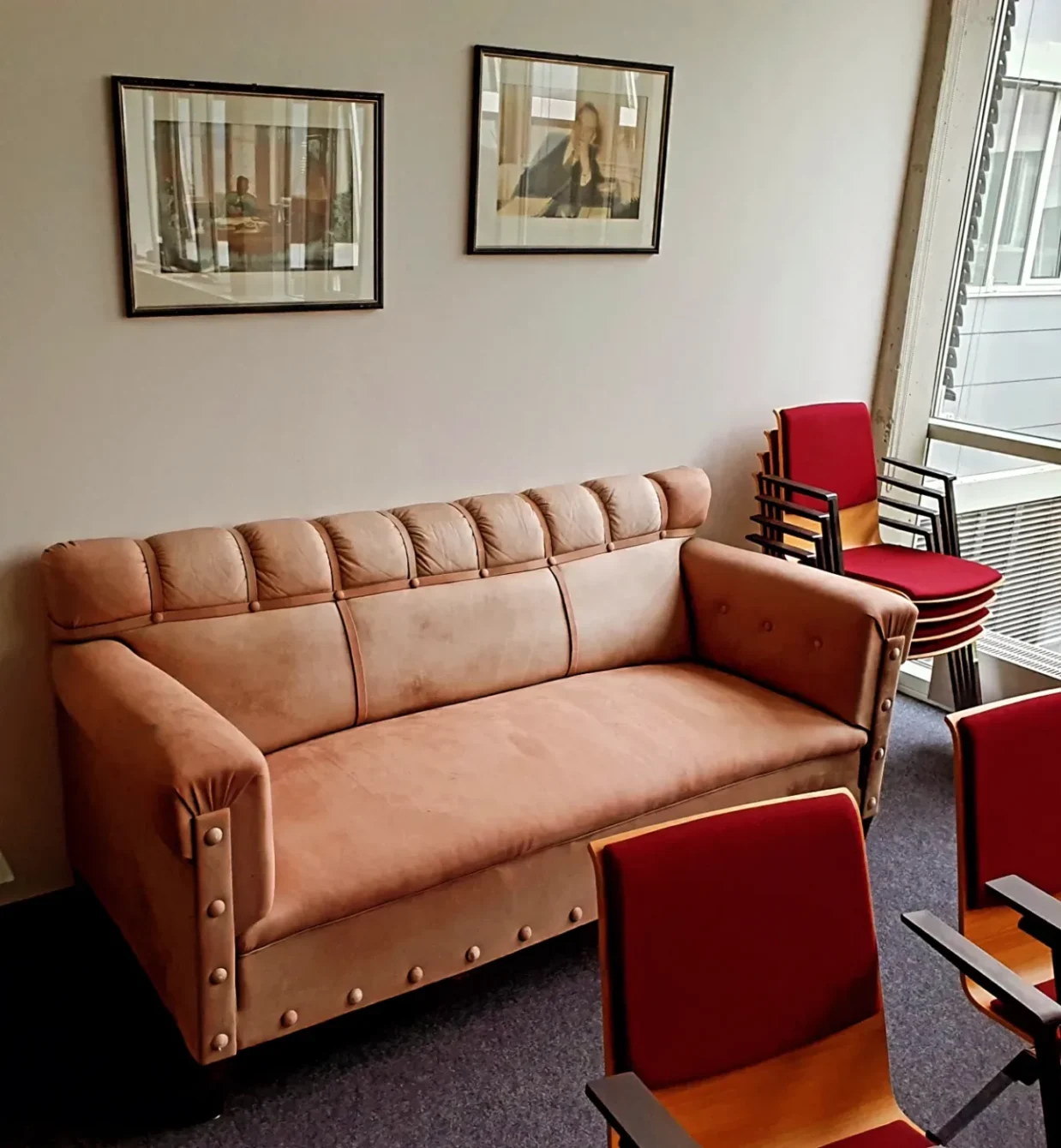 A retro tan leather sofa next to red stackable chairs, and framed artwork on the wall. The space has a vintage office feel, with a window visible on the right side.