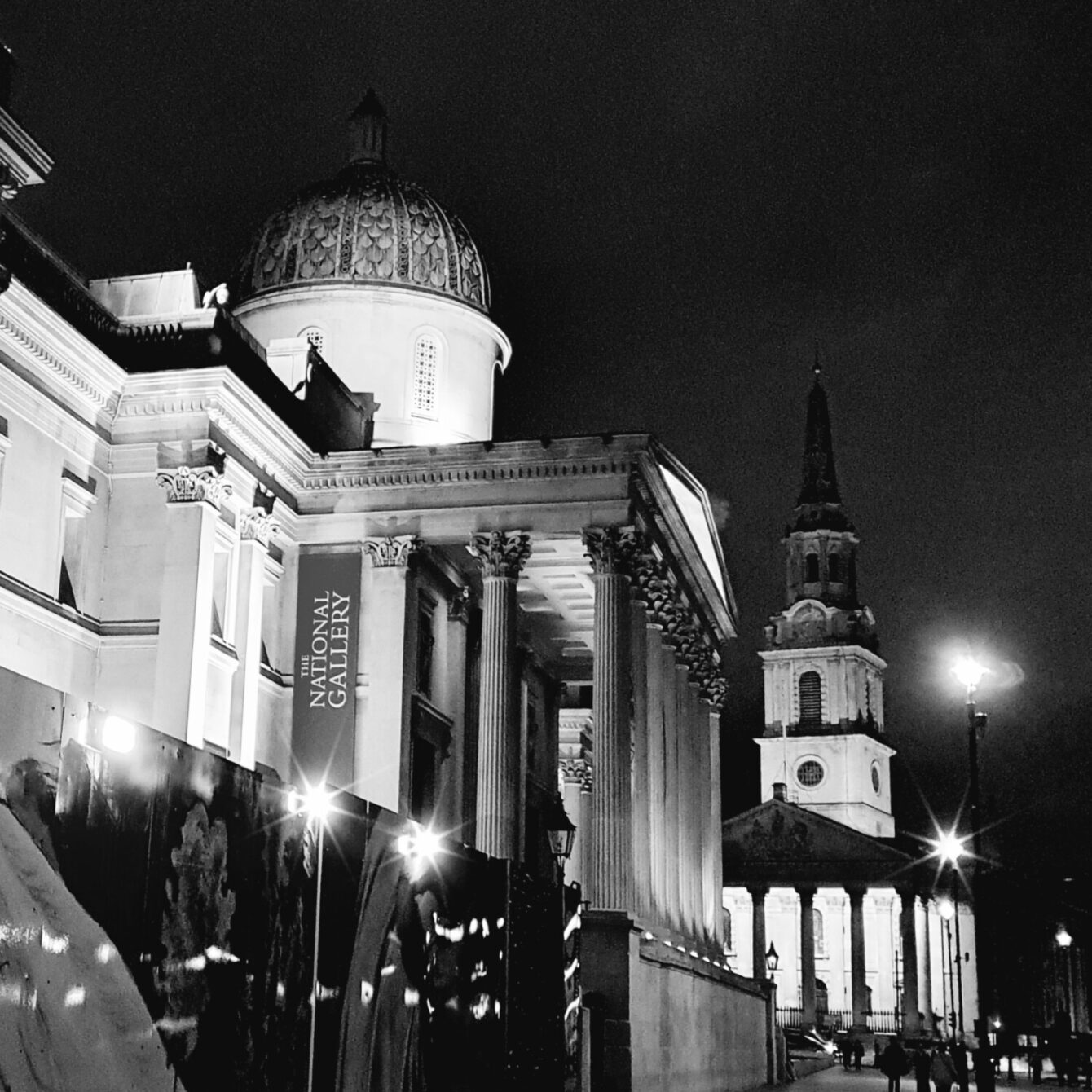 Night view of London's National Gallery and St. Martin-in-the-Fields church. The neoclassical facade of the gallery with its iconic dome is illuminated, contrasting with the dark sky. Street lamps create a warm glow in this black and white urban scene.