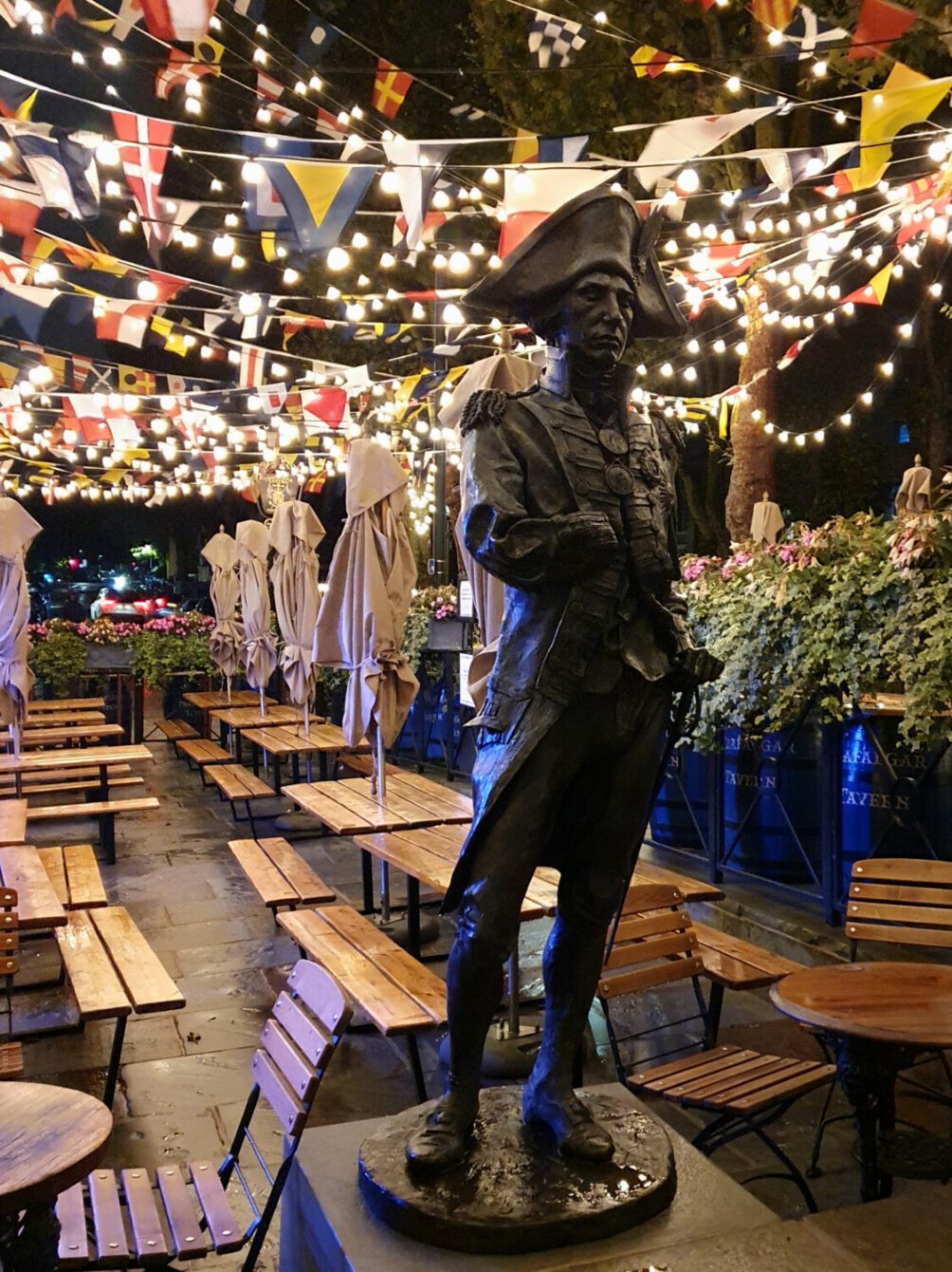 Outdoor pub garden with statue of naval officer, festive string lights, and international flags creating a cozy ambiance.