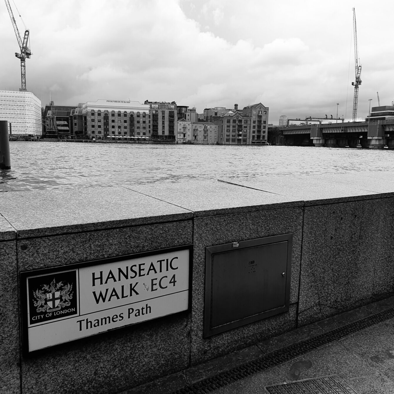 Black and white image of Thames riverfront in London, featuring Hanseatic Walk sign, warehouses, and construction cranes against a cloudy sky.