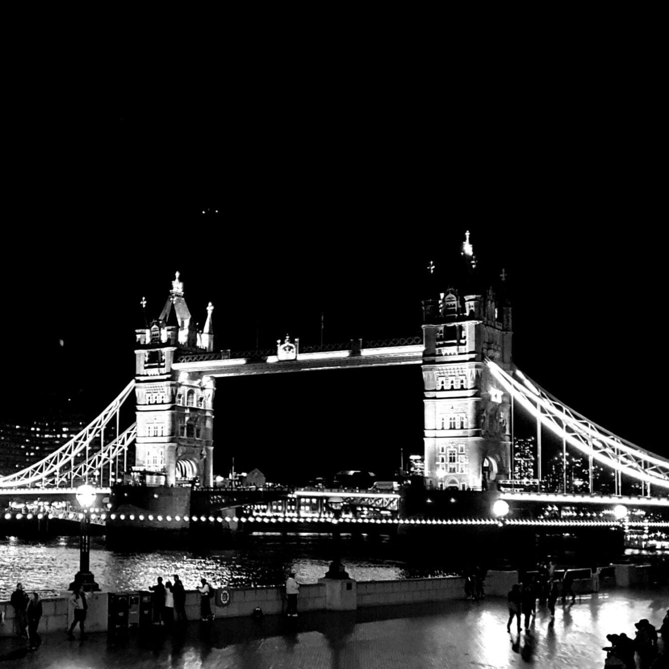 Tower Bridge illuminated at night, London skyline. Black and white image shows iconic bridge with tourists on riverside walkway.