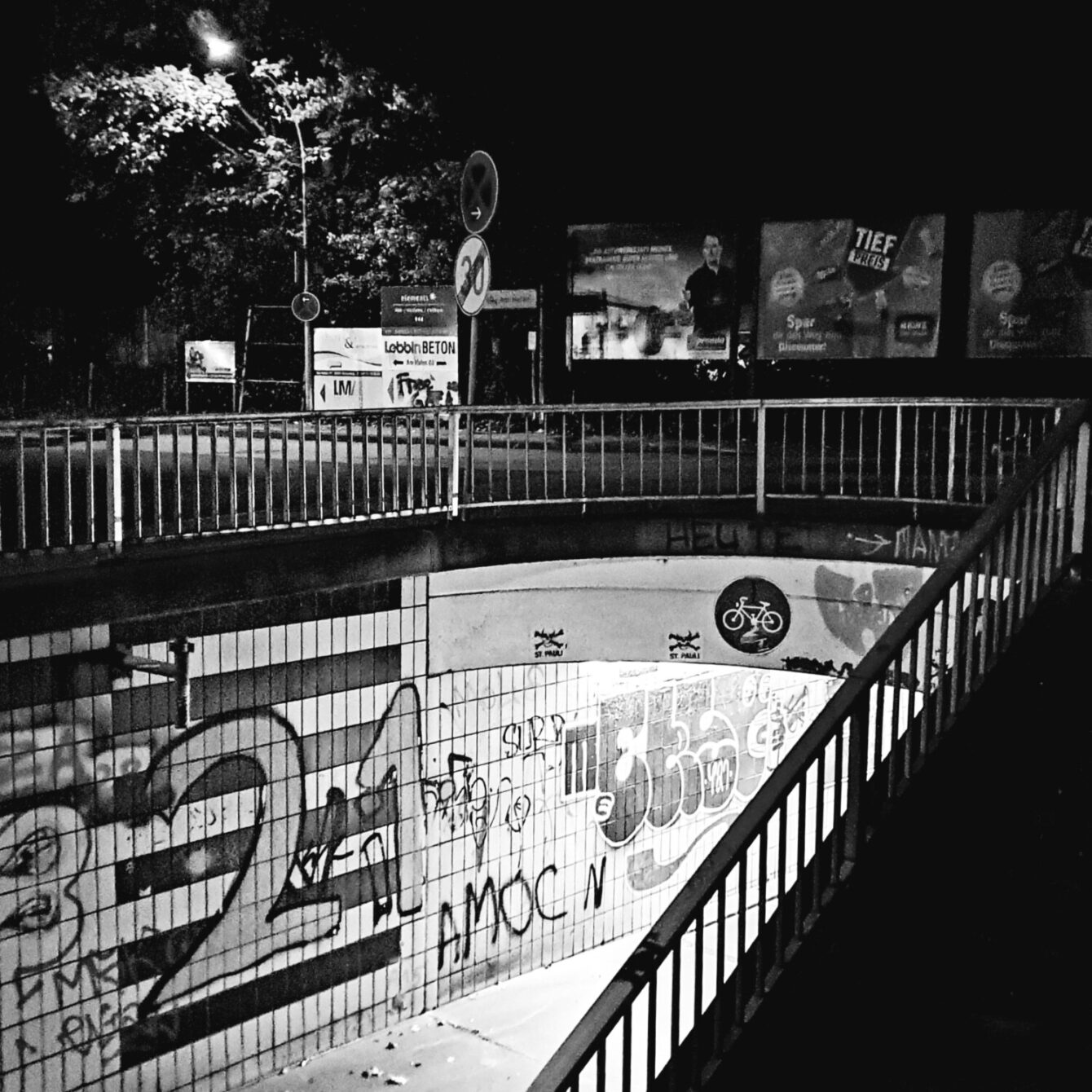 Black and white night shot of an urban underpass with graffiti, bicycle sign, and metal railings. Street art and advertising boards visible in the background.