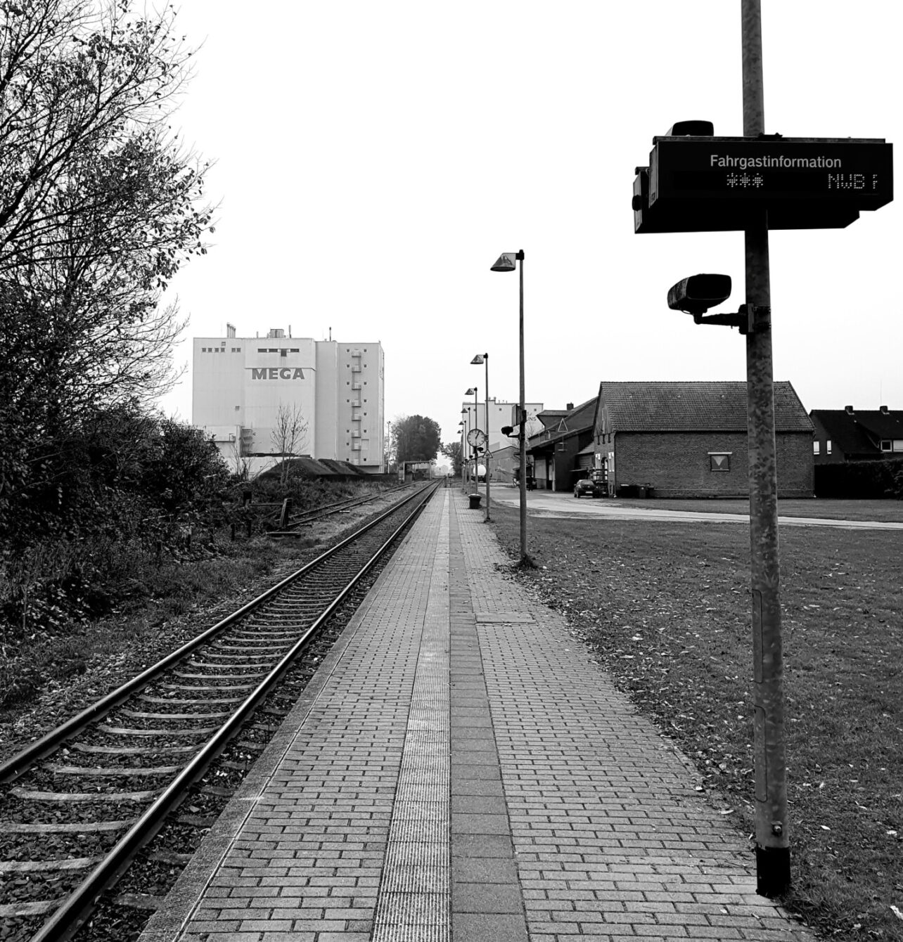 Black and white photo of a German train platform with building in background. Electronic passenger information display and railway tracks leading to horizon.