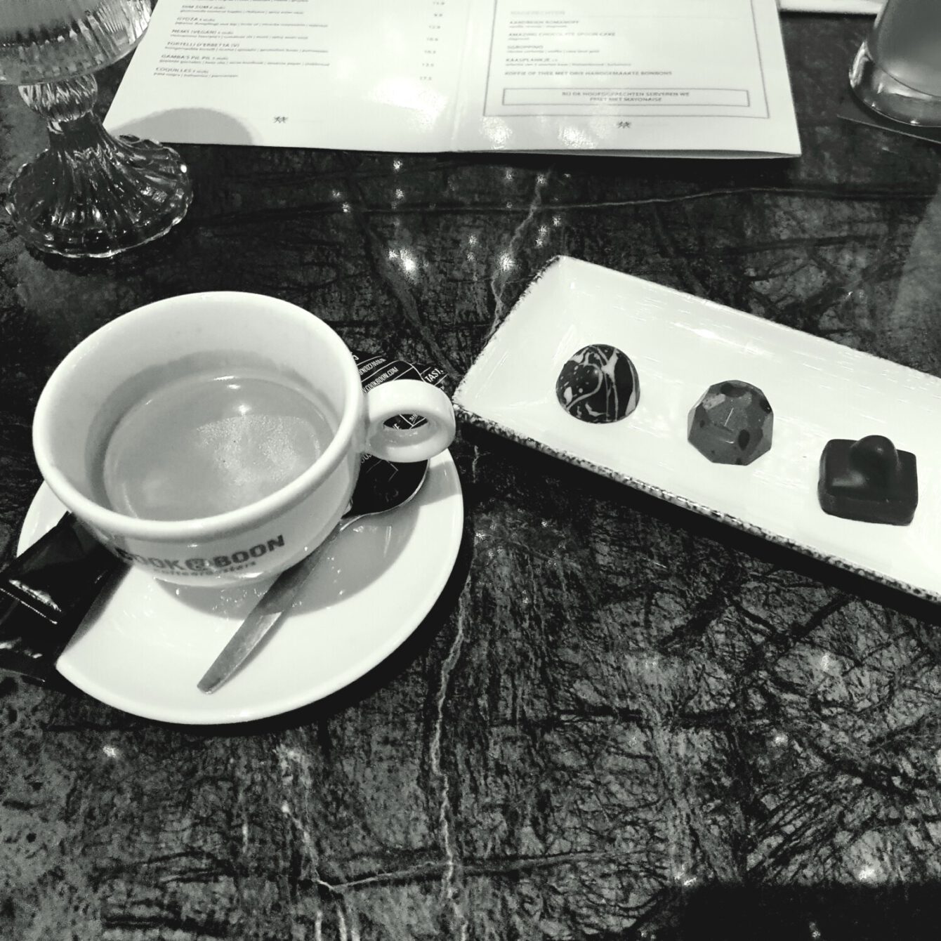 Black and white photo of a coffee cup with artisanal chocolates served on a marble table, restaurant menu visible.