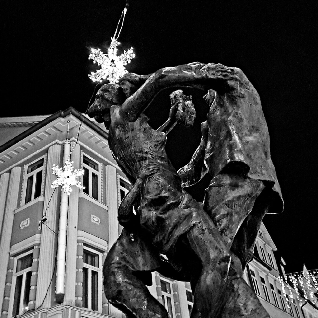 Black and white nighttime photograph of a dynamic bronze sculpture showing two dancing figures, set against a historic building facade decorated with illuminated snowflake ornaments. The contrast between the dark sculpture and the lit architectural elements creates a dramatic winter atmosphere.