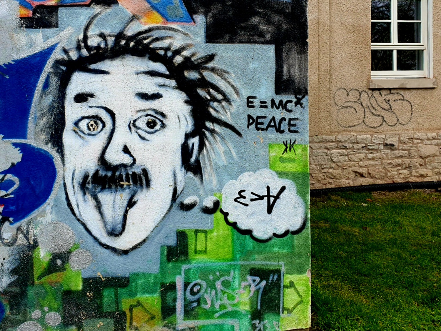 Colorful street art mural featuring a portrait of Einstein sticking out his tongue, surrounded by bright blue and green graffiti elements, with 'PEACE' written nearby. The artwork combines playful and scientific themes in an urban style.