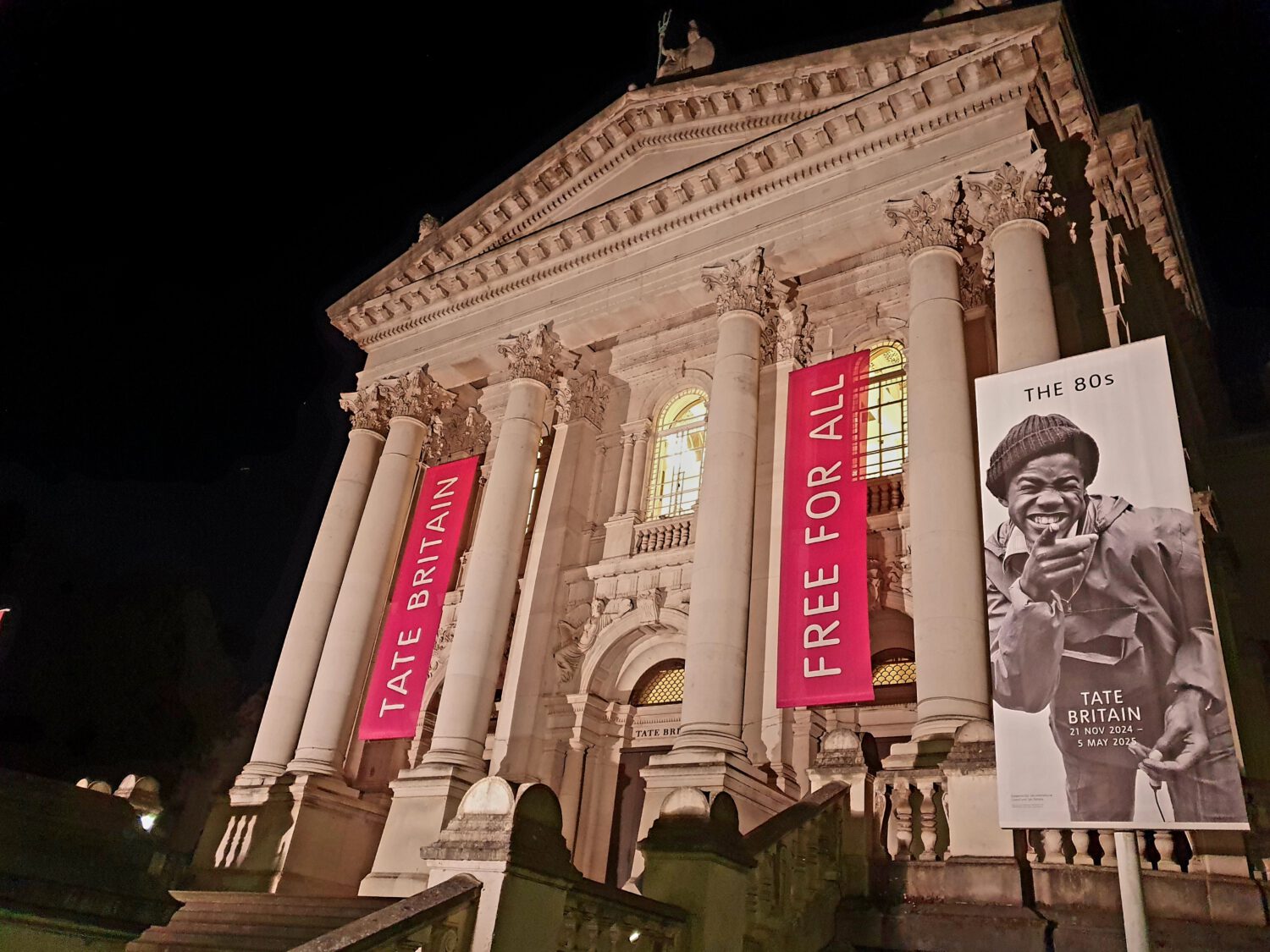 Tate Britain museum illuminated at night with bright pink banners reading 'FREE FOR ALL' and a large poster for 'THE 80s' exhibition running from 21 November 2024 to 5 May 2025. The neoclassical facade features ornate columns and decorative details.