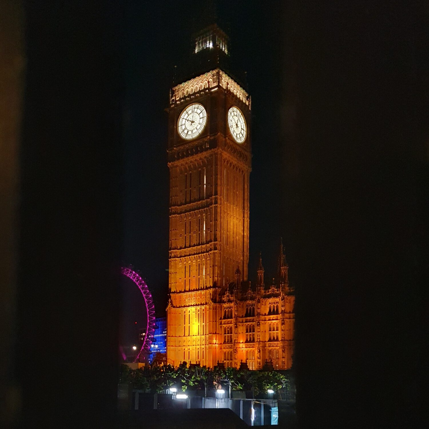 Elizabeth Tower (Big Ben) illuminated in golden light against the night sky, with its iconic clock faces glowing. The Houses of Parliament building is visible at the base, and the colourful London Eye can be spotted in the background with purple illumination.