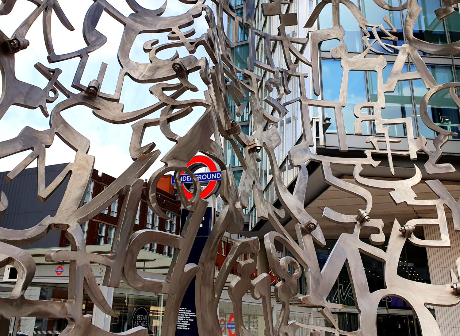 Abstract metal sculpture with letter-like forms in London, framing a London Underground roundel sign. The modern sculptural installation creates an artistic gateway near a glass building, showcasing the blend of contemporary art and iconic city transport branding.