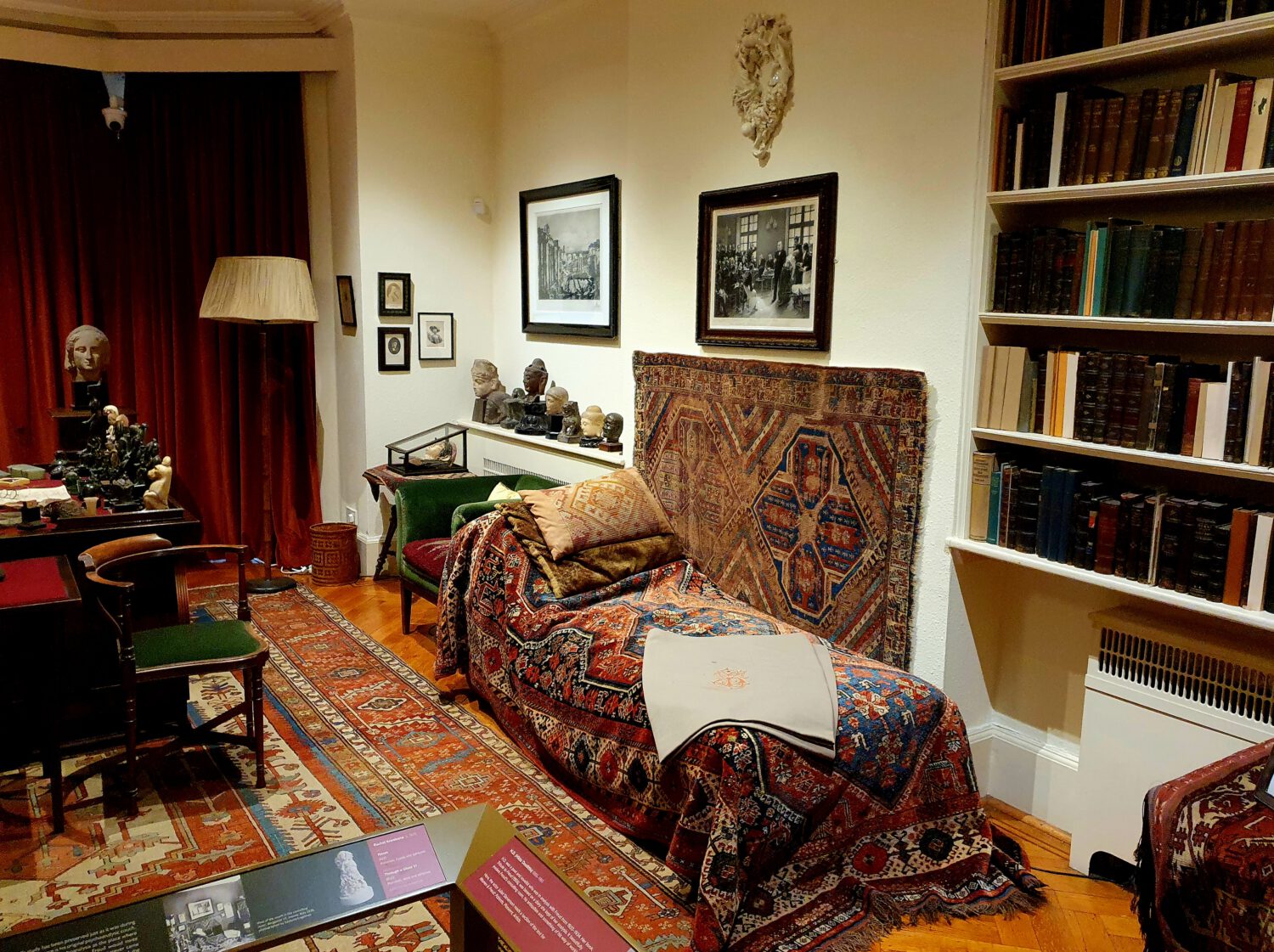 Cozy historic study room with vintage furnishings including oriental rugs, a chaise lounge covered in patterned fabrics, built-in bookshelves, framed artworks, and decorative busts. The warm, scholarly atmosphere suggests a preserved historic home or museum space.