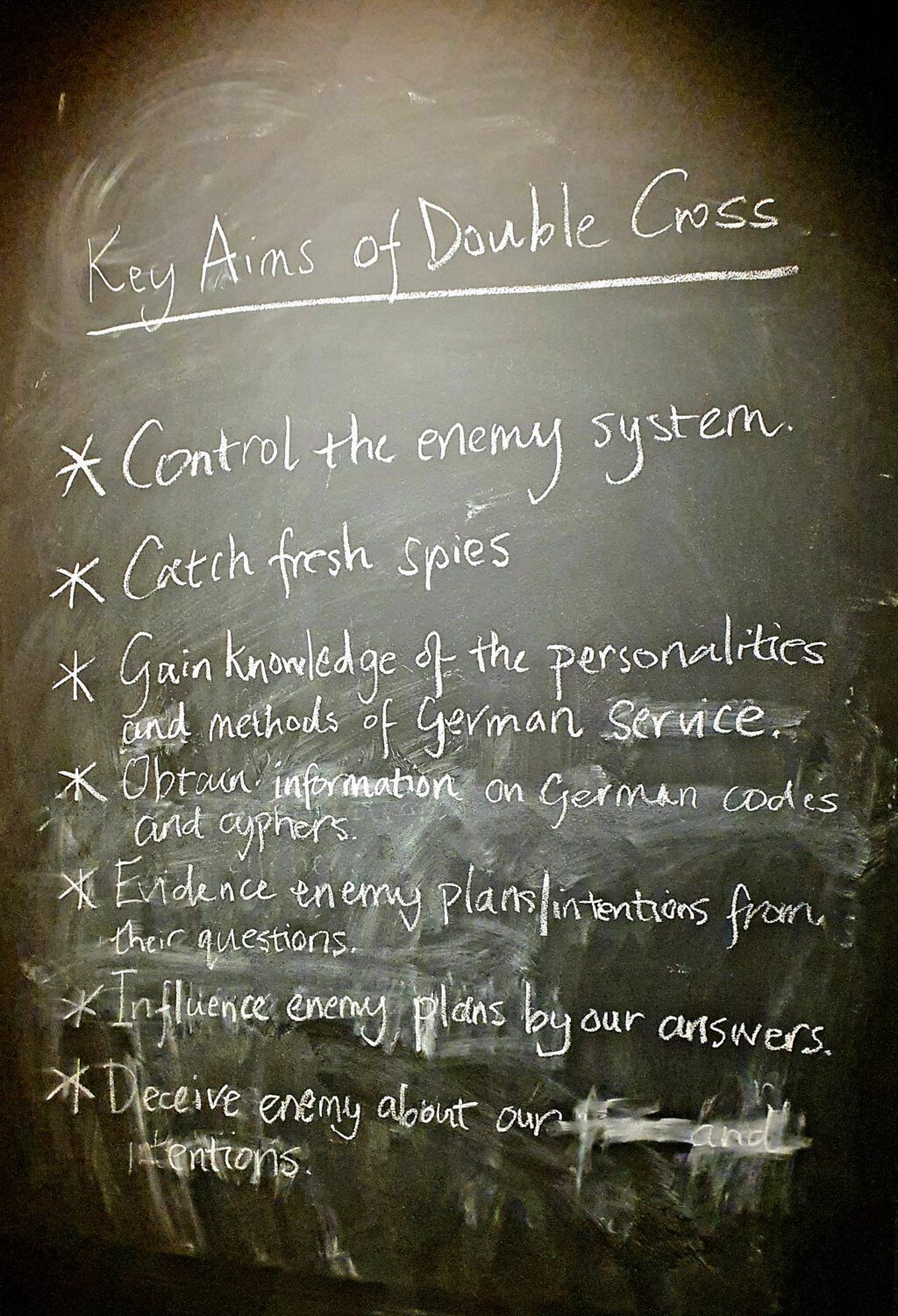 Handwritten chalk board listing 'Key Aims of Double Cross' with seven strategic points about intelligence operations during wartime, including controlling enemy systems, catching spies, and gathering information on German codes and ciphers. Historical museum display of WWII espionage tactics.