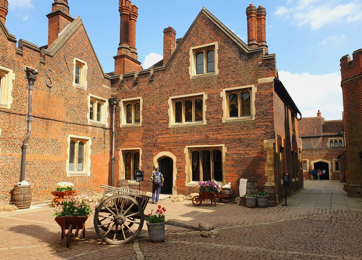 Tudor-style historic brick building with distinctive chimney stacks and mullioned windows. The courtyard features wooden carts with spring flowers, traditional barrels, and cobblestone paving. A visitor explores this well-preserved heritage site on a sunny day.