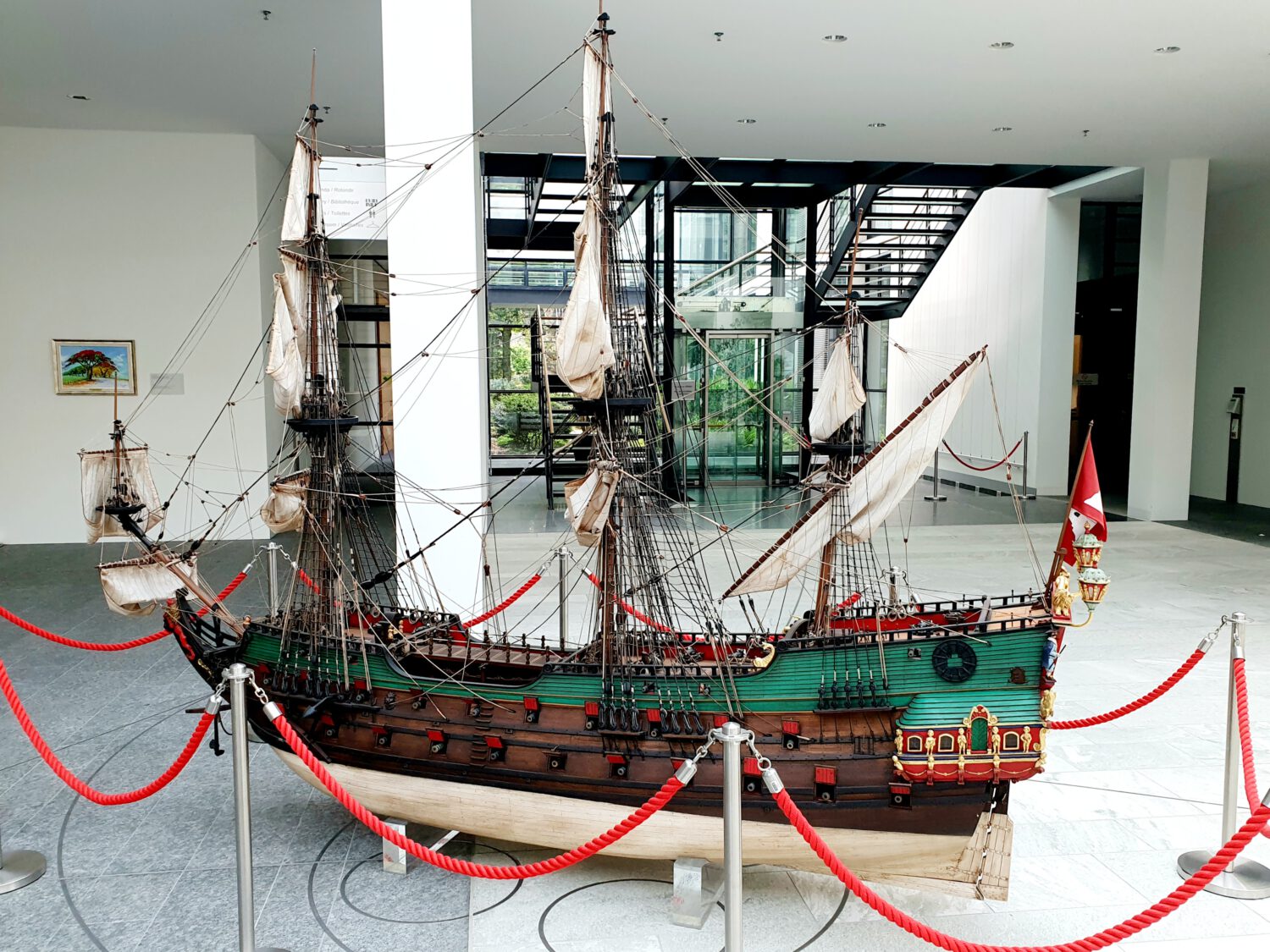 Large model sailing ship with green hull and white sails displayed in a modern museum gallery. The detailed replica features multiple masts with rigging and decorative stern elements. Red rope barriers on metal posts surround the exhibit. Modern architecture visible in background includes stairs, large windows, and white walls with contemporary lighting.