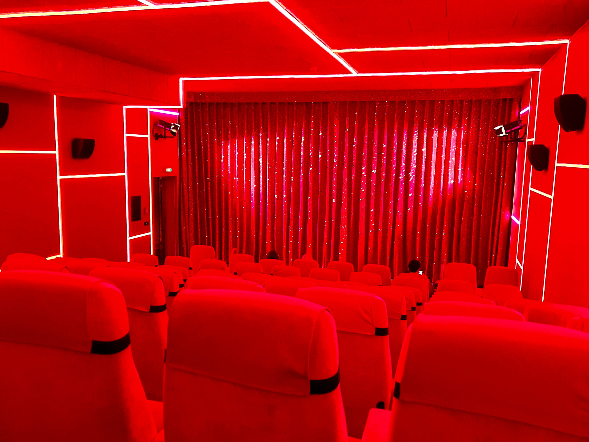 Interior of a small cinema bathed entirely in vibrant red lighting. Red theater seats face a stage with closed red curtains, while geometric white light fixtures create angular patterns on the red walls and ceiling. The monochromatic red colour scheme creates a dramatic, immersive atmosphere.
