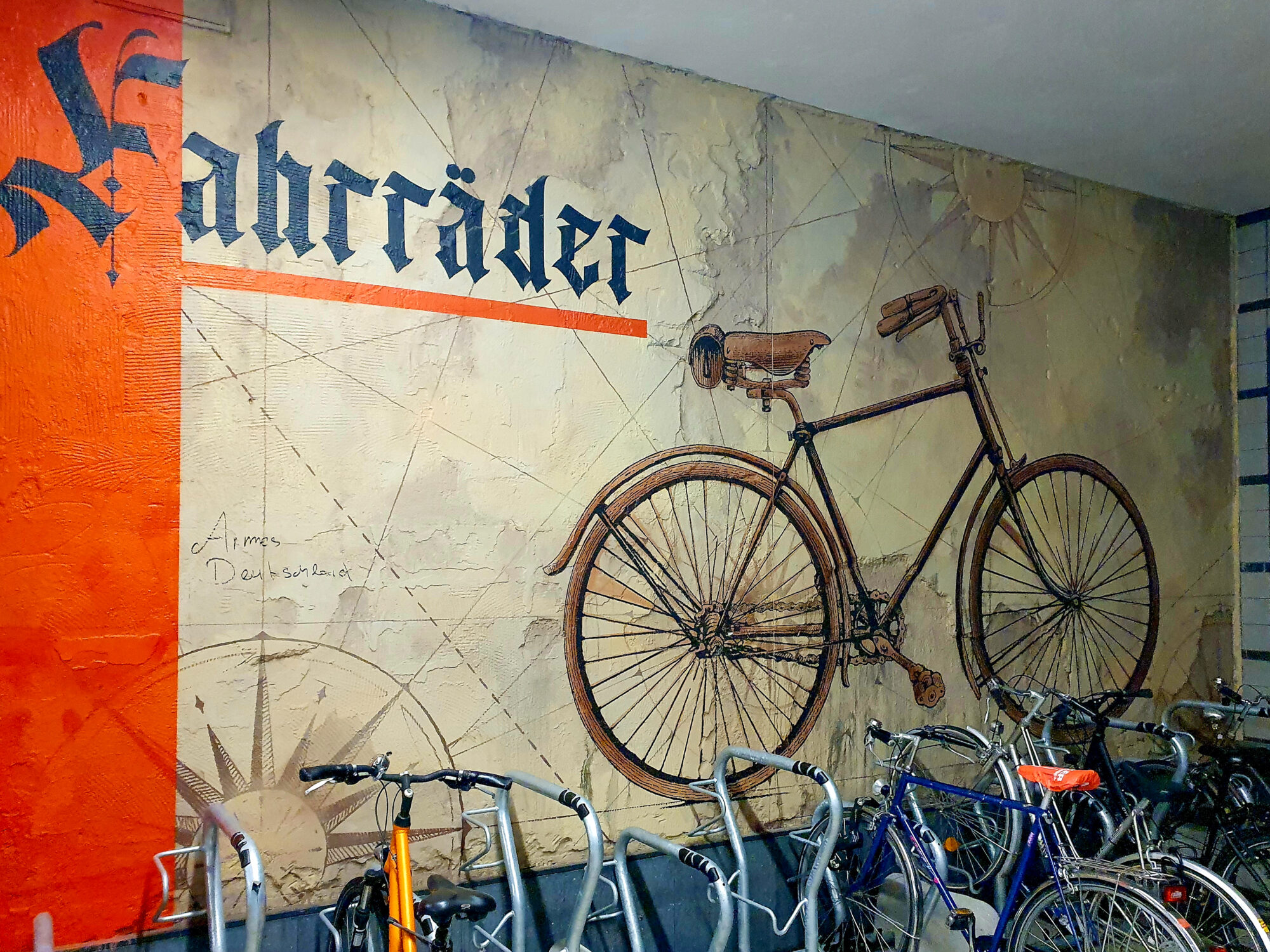 Wall mural in a bicycle parking area featuring a vintage bicycle design and Gothic lettering reading "Fahrräder." The artwork shows a classic brown bicycle painted on a cream-colored wall with an orange accent stripe. Modern bicycles are parked below the mural in metal racks.