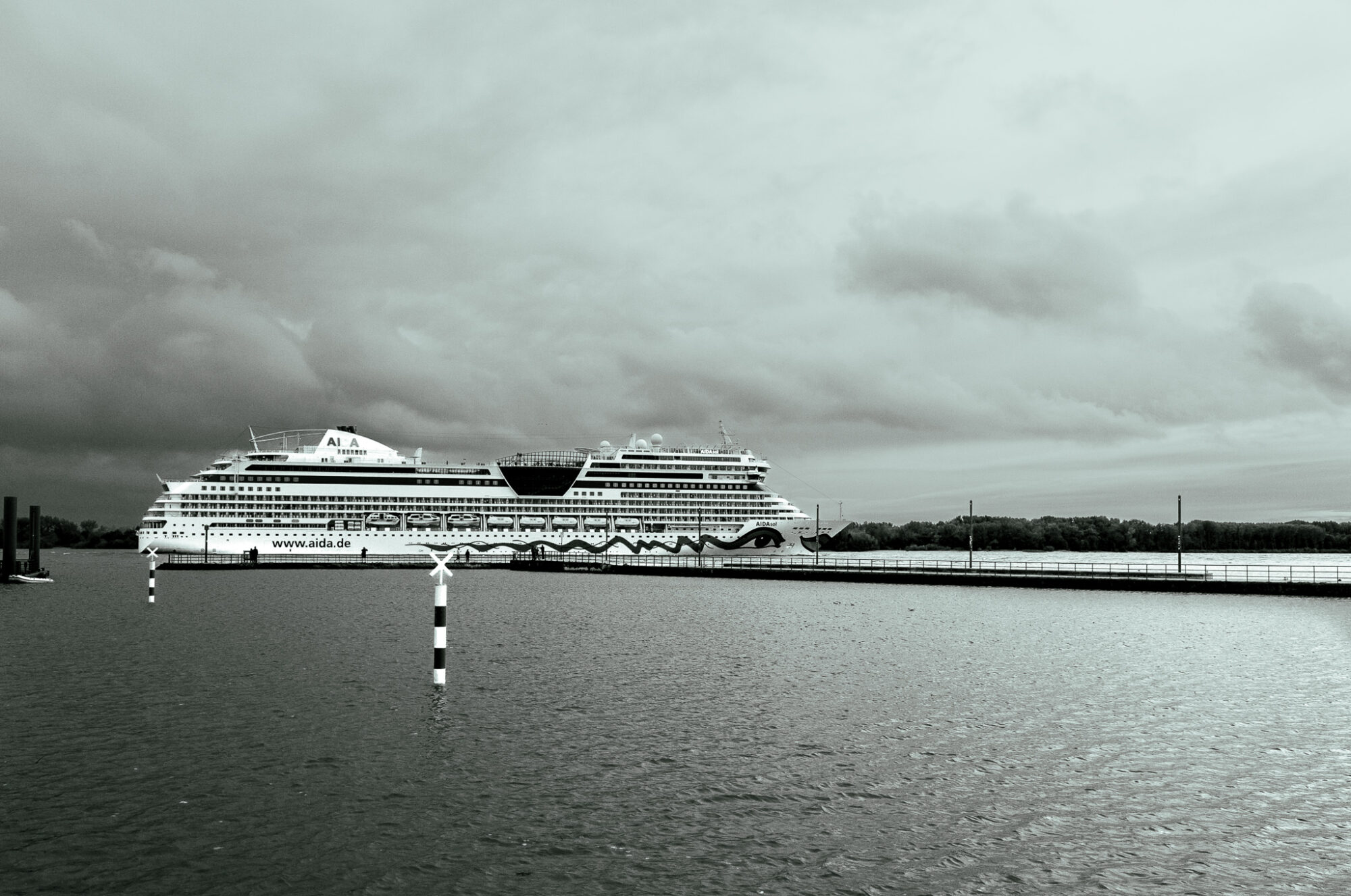 Large white cruise ship with "AIDA" branding passing by a pier under an overcast sky. A striped navigation marker pole stands in the foreground water, with the ship's multiple decks and distinctive funnel design visible against grey clouds.