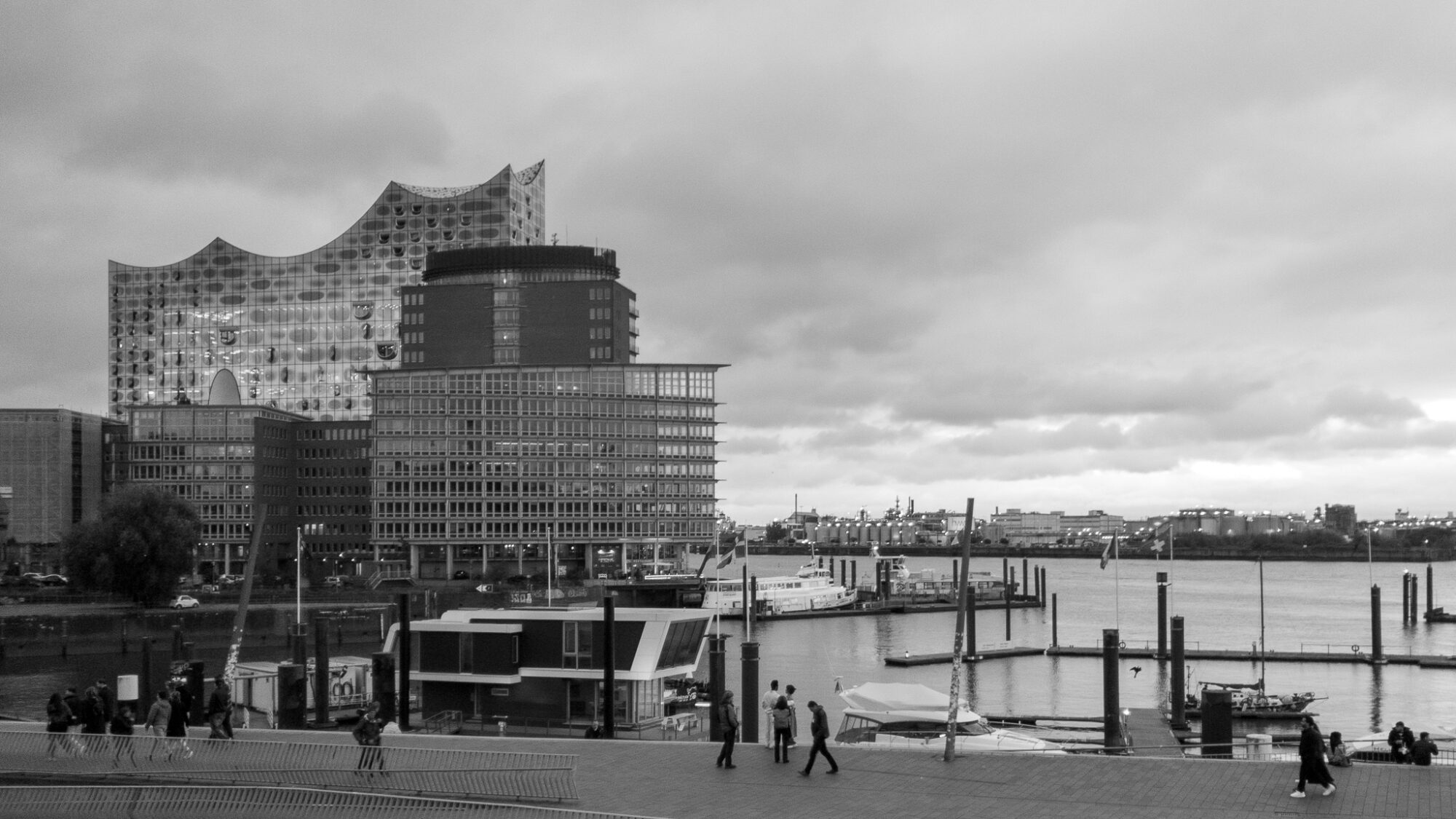 Black and white photograph of Hamburg's Elbphilharmonie concert hall rising above the harbour. The distinctive wave-shaped glass structure sits atop a historic brick warehouse building. In the foreground, pedestrians walk along the waterfront promenade near docked boats and marina pilings. Office buildings line the opposite shore under an overcast sky.
