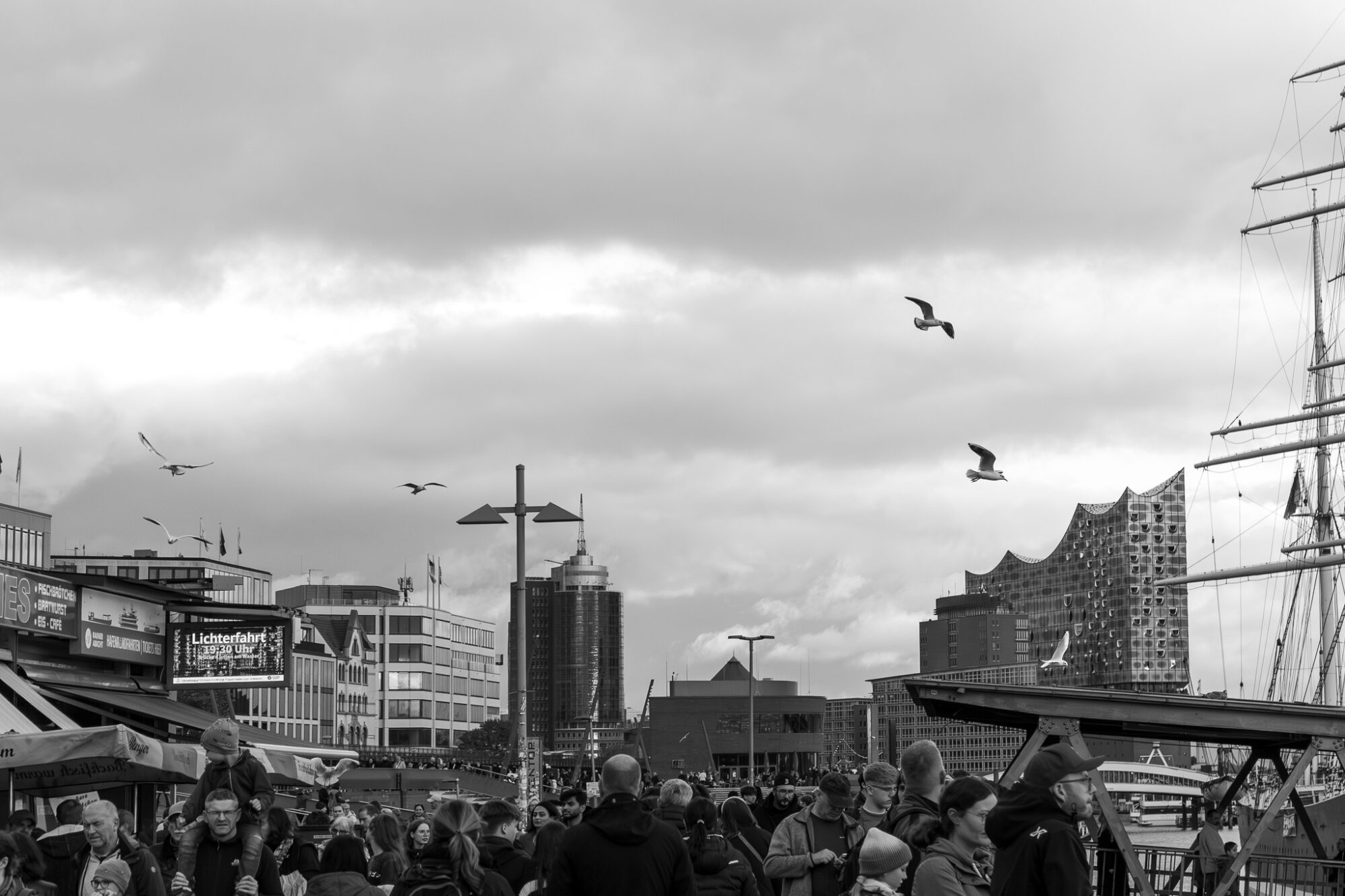 Black and white photograph of a crowded waterfront promenade with seagulls flying overhead against a cloudy sky. The Elbphilharmonie's distinctive wave-like architecture dominates the right side of the frame, while a tall ship's mast is visible at the far right. Various modern and historic buildings line the harbour, with pedestrians filling the walkway below.