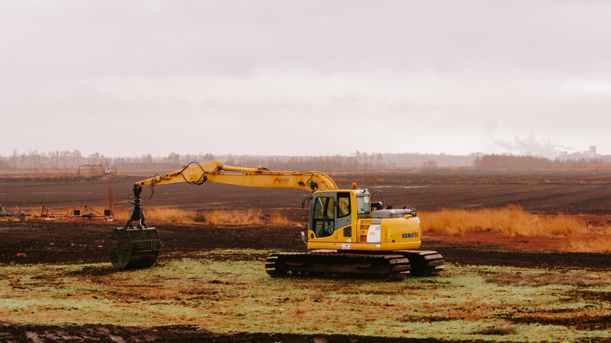 Yellow excavator with an extended arm and grapple attachment sits on burnt moorland. The machine appears to be involved in peat extraction or restoration work, with golden dried grasses in the foreground and a hazy horizon.