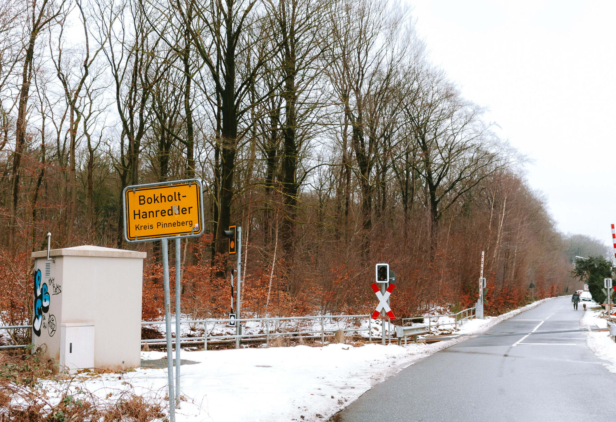 A rural winter scene showing a railway crossing at Bokholt-Hanredder in Kreis Pinneberg, Northern Germany. The yellow German town sign stands beside a graffitied utility box on the left, while bare deciduous trees with remaining brown leaves create a backdrop against a misty sky. Light snow covers the ground along the asphalt road, which extends into the distance where a cyclist can be seen. Red and white railway crossing barriers and signals are visible in the midground.