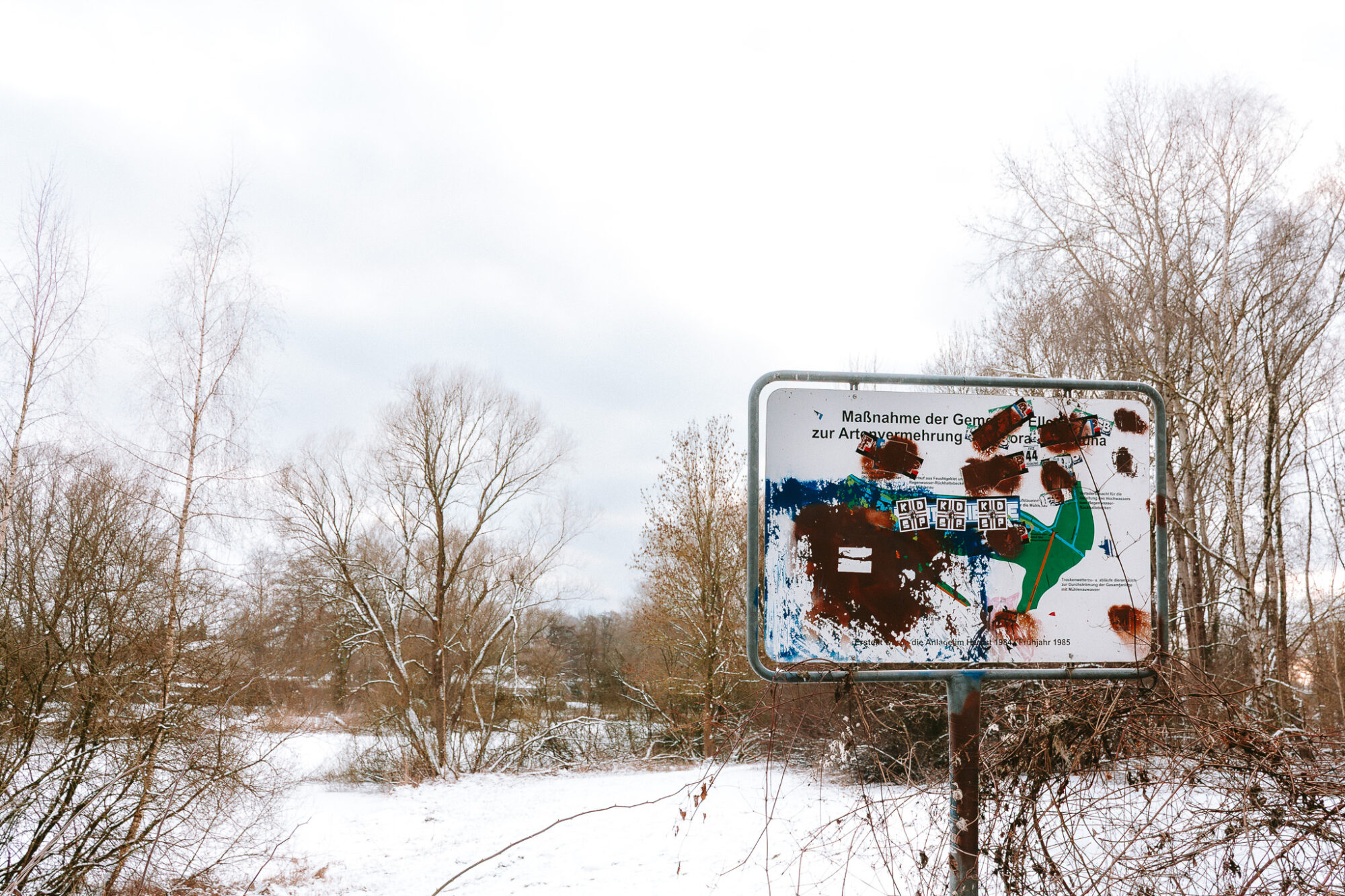 A weathered information sign stands in a winter landscape, its surface heavily damaged with peeling paint and rust stains. The sign appears to show a map with text in German and colourful graphics, but much of it is obscured by deterioration. Behind it stretches a snow-dusted field with bare trees and a grey overcast sky, creating a melancholic atmosphere of abandonment.