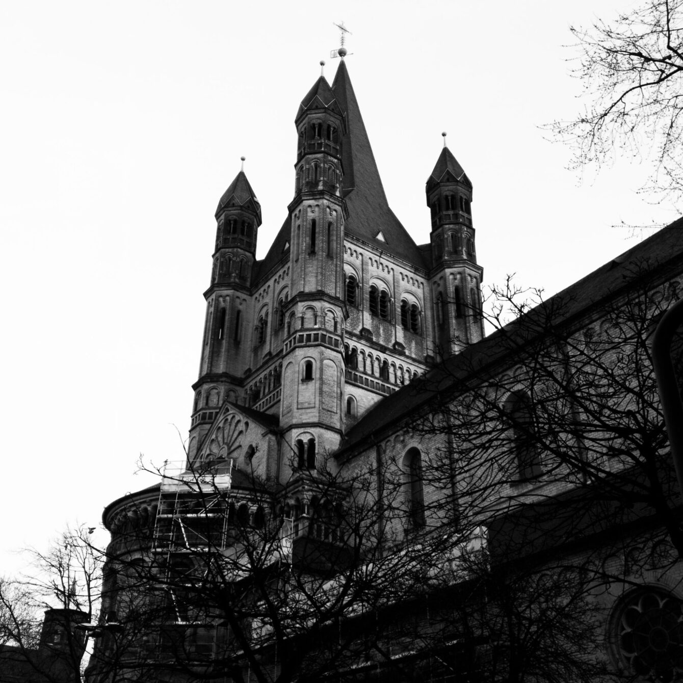 A black-and-white upward-looking photograph of a large Romanesque church tower complex, with a tall central spire flanked by four smaller turrets. Ornate arcading and rounded arches decorate the tower. Bare winter branches reach into the frame from the right, and scaffolding is visible at the lower left.