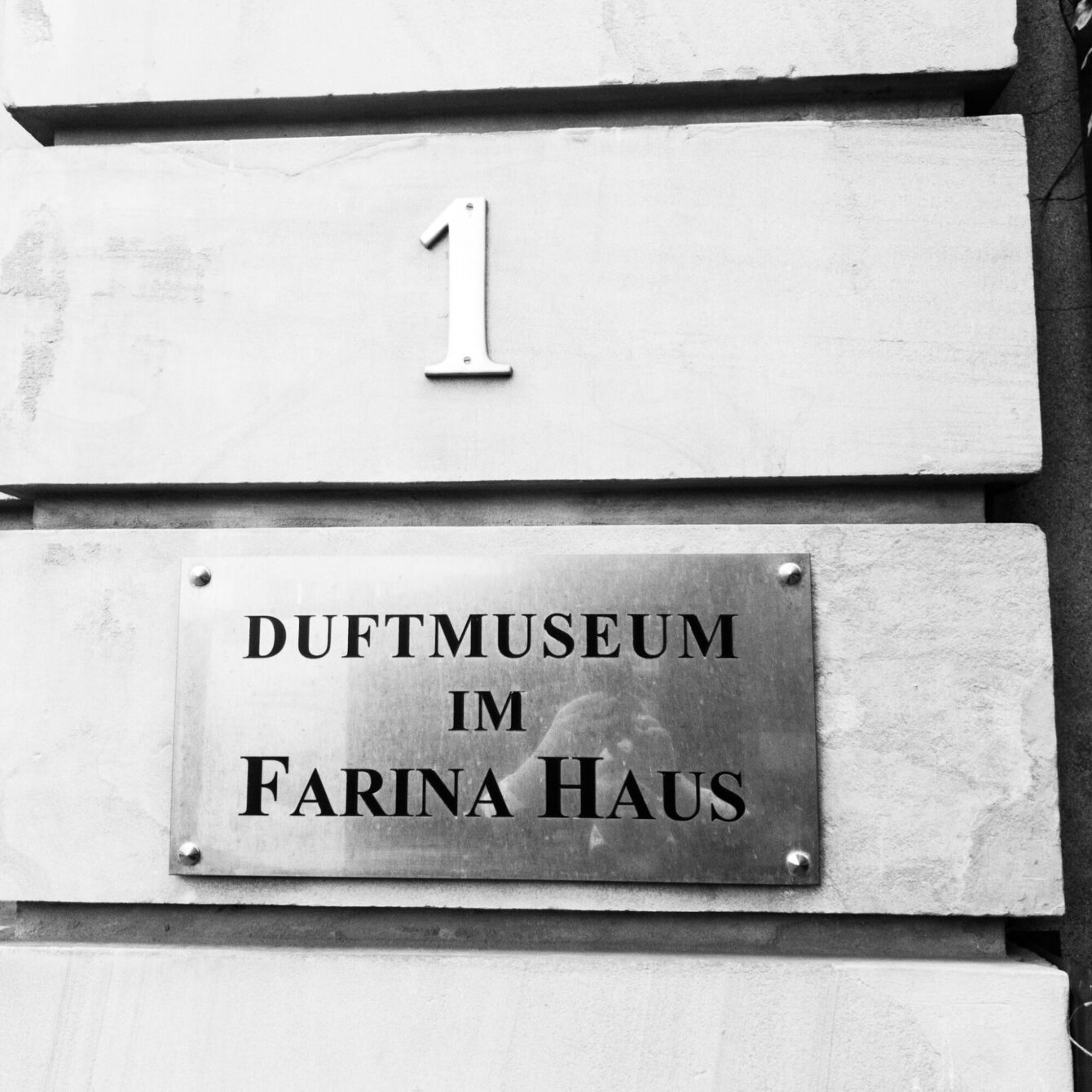 A close-up black-and-white photograph of a stone façade with a polished metal plaque reading "Duftmuseum im Farina Haus" and the house number 1 mounted above it. The photographer's reflection is faintly visible in the metal surface of the plaque.