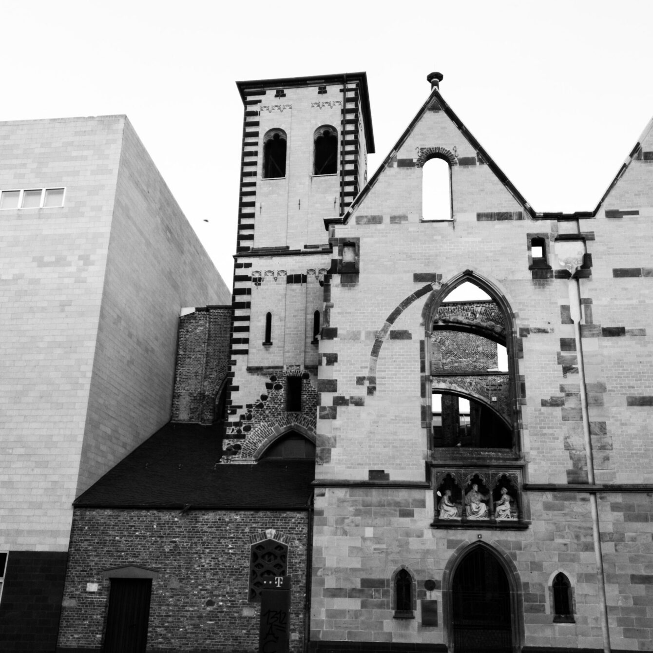 A black-and-white low-angle photograph of a partially ruined medieval church, its stone walls open to the sky where windows once were. A slender tower with alternating light and dark stonework rises on the left, and gothic sculptural reliefs are visible above a pointed arch. A plain modern building stands immediately adjacent on the left.