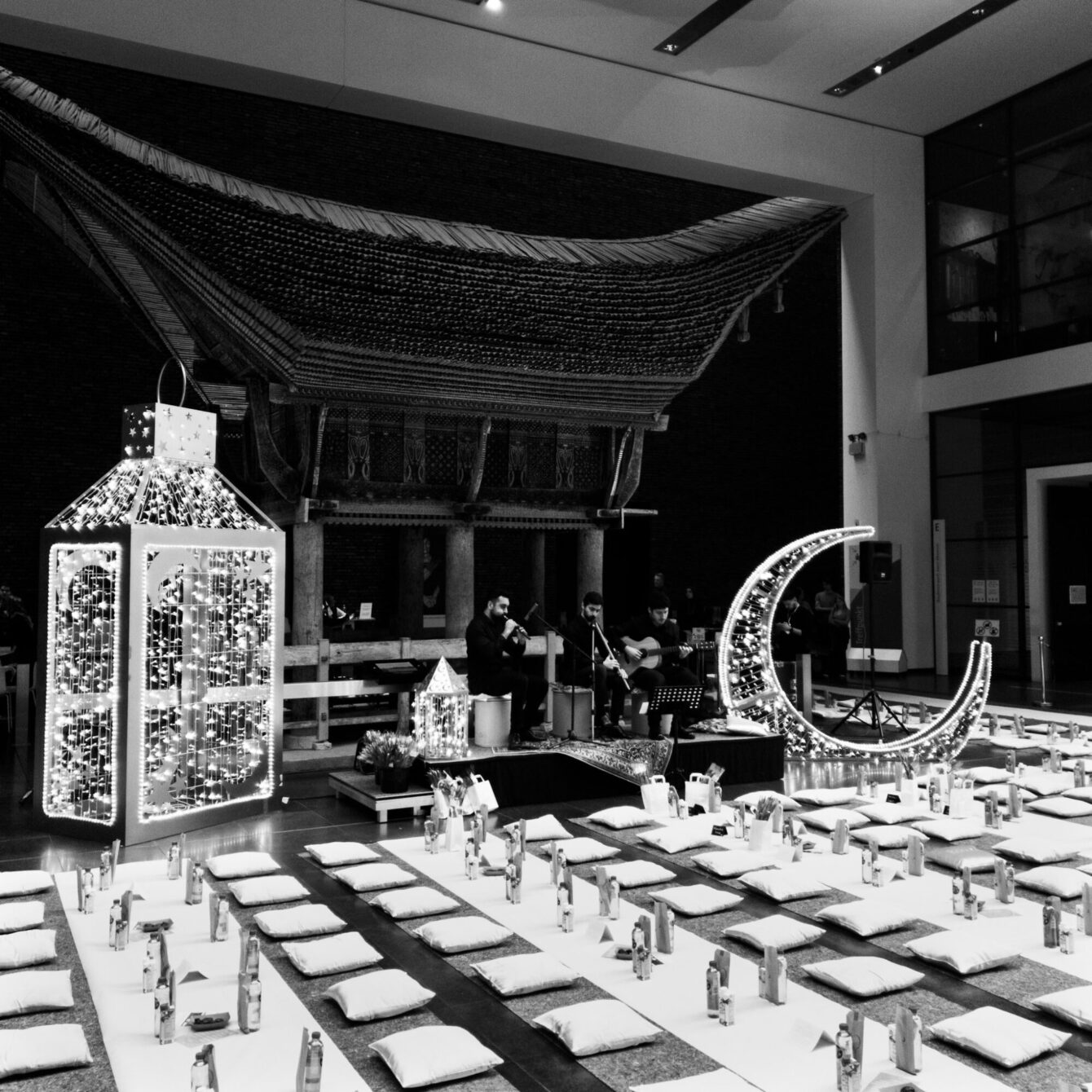 A black-and-white interior shot of a large museum hall set up for an evening event. Rows of flat cushions and small candles are arranged on the floor facing a raised stage. Behind the performers — one playing violin, one guitar — stands a traditional carved wooden Toraja house from Sulawesi. To the left, a tall illuminated lantern structure; to the right, a glowing crescent moon decoration.