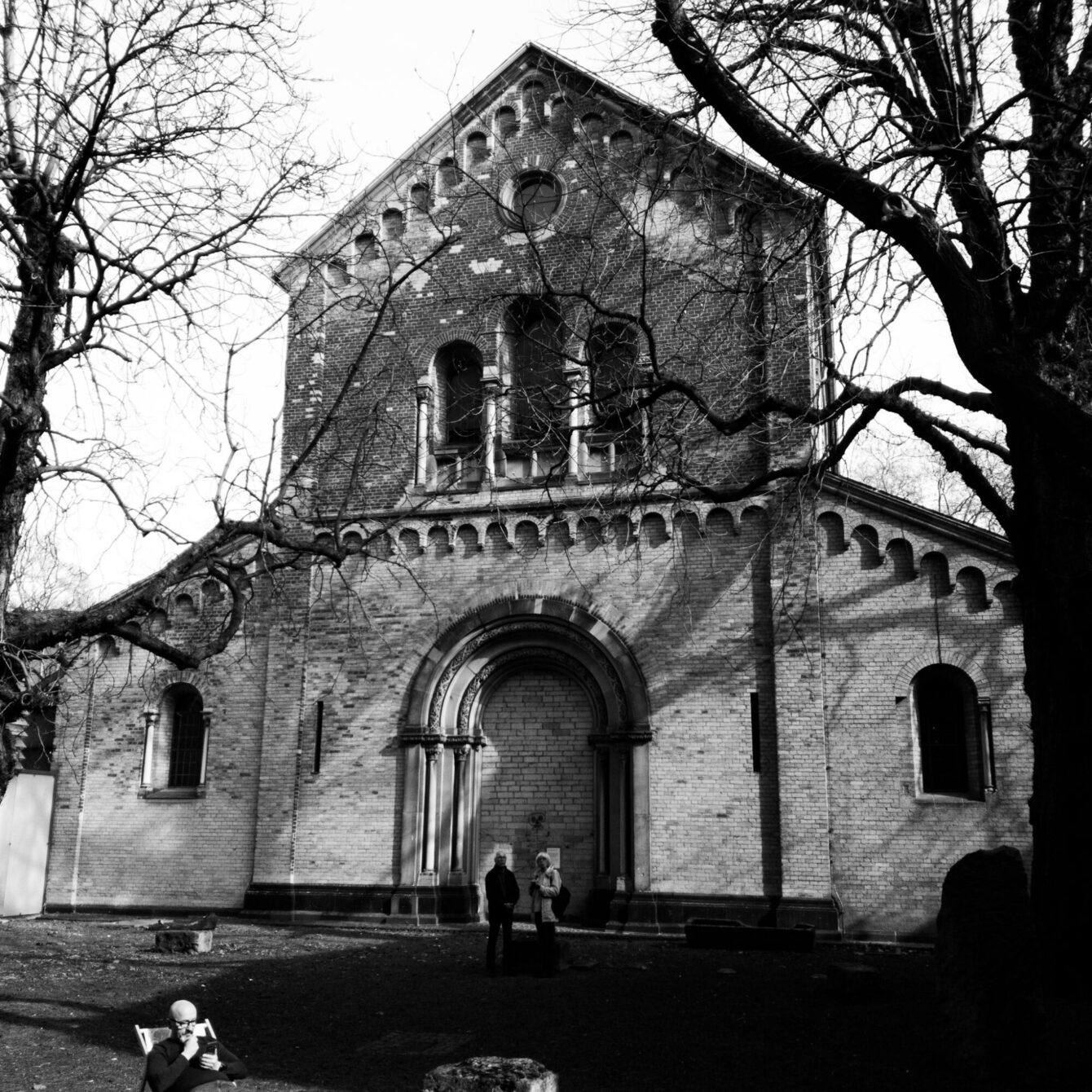 A black-and-white photograph of a Romanesque brick church façade, featuring a wide arched entrance portal, a row of small blind arches along the base, and a gabled upper section with circular and arched windows. Bare winter trees partially obscure the building. Two people stand at the entrance, and a seated figure reads in the foreground.