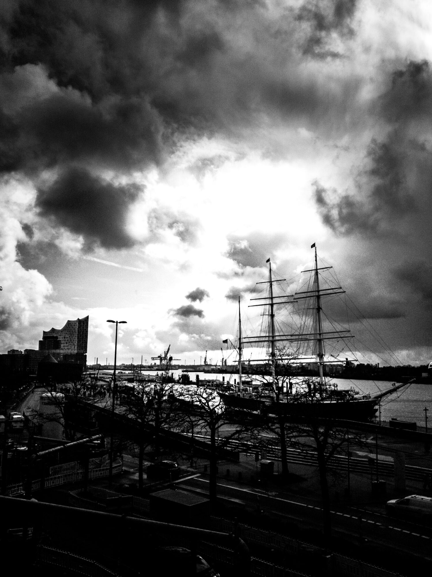 Black and white photograph of Hamburg's harbour waterfront. A tall sailing ship with multiple masts and rigging dominates the right side, moored at the dock. On the left horizon, the distinctive wave-shaped façade of the Elbphilharmonie concert hall is visible as a silhouette. Industrial port cranes stretch across the middle distance. Bare winter trees and a busy quayside promenade fill the foreground. Dramatic storm clouds billow across a bright sky, creating strong contrast in this moody cityscape.