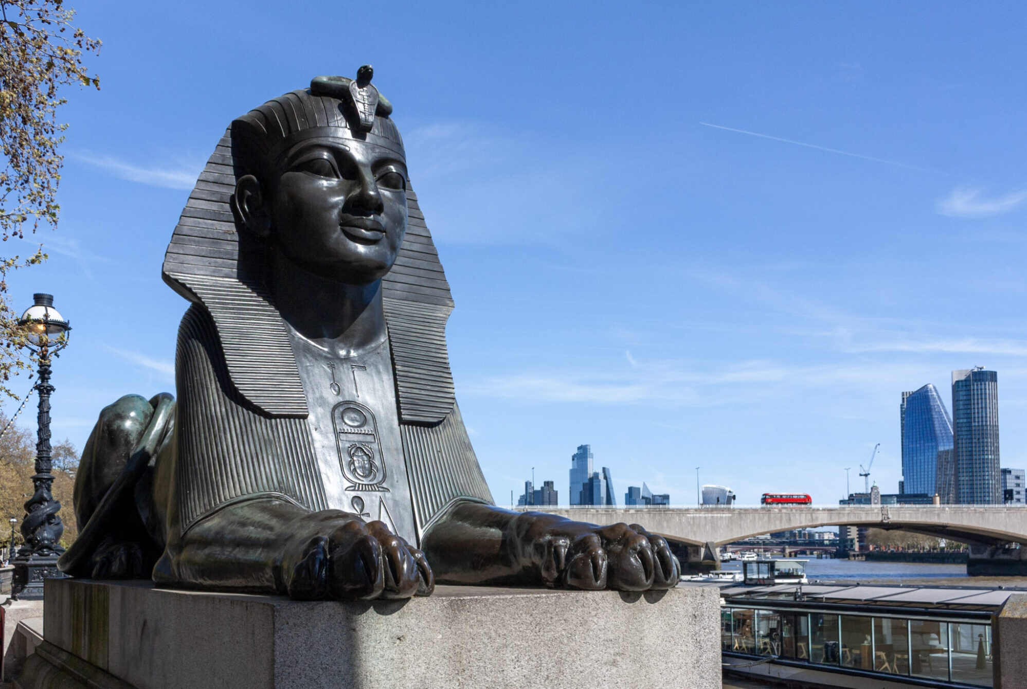 A dark bronze sphinx statue on the Victoria Embankment in London sits in the foreground, its lion paws extended forward on a stone plinth. Egyptian hieroglyphics and a cartouche are visible on its chest. Behind it, the Thames stretches toward Waterloo Bridge, with a red double-decker bus crossing. The South Bank skyline — including the distinctive curved Vase building and tall glass towers — rises under a clear blue spring sky. An ornate Victorian lamp post stands to the left beside bare-branched trees.