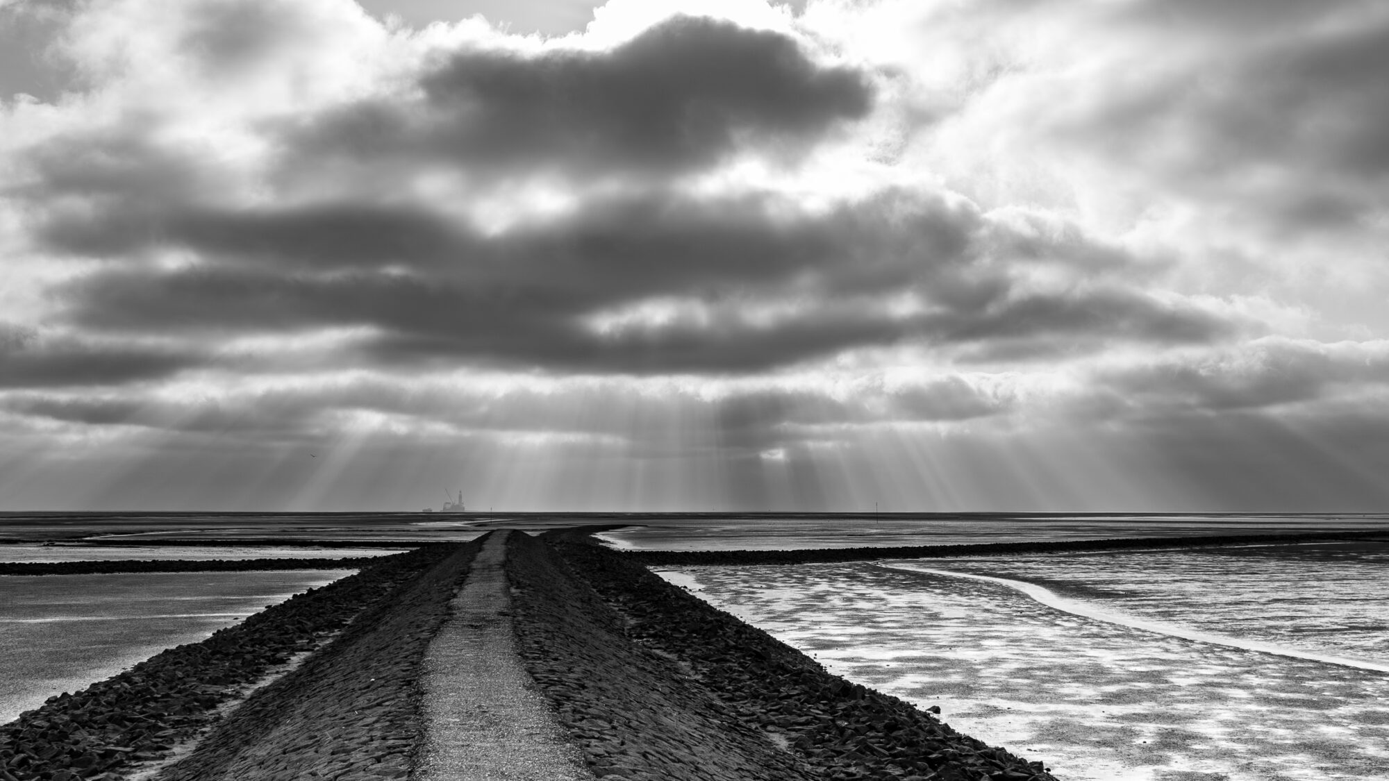 Black-and-white photograph of a raised causeway leading straight towards the horizon across the Wadden Sea flats, flanked by rocky embankments and shallow pools. Dramatic crepuscular rays break through heavy clouds above, and the faint silhouette of an offshore platform is visible in the distance.