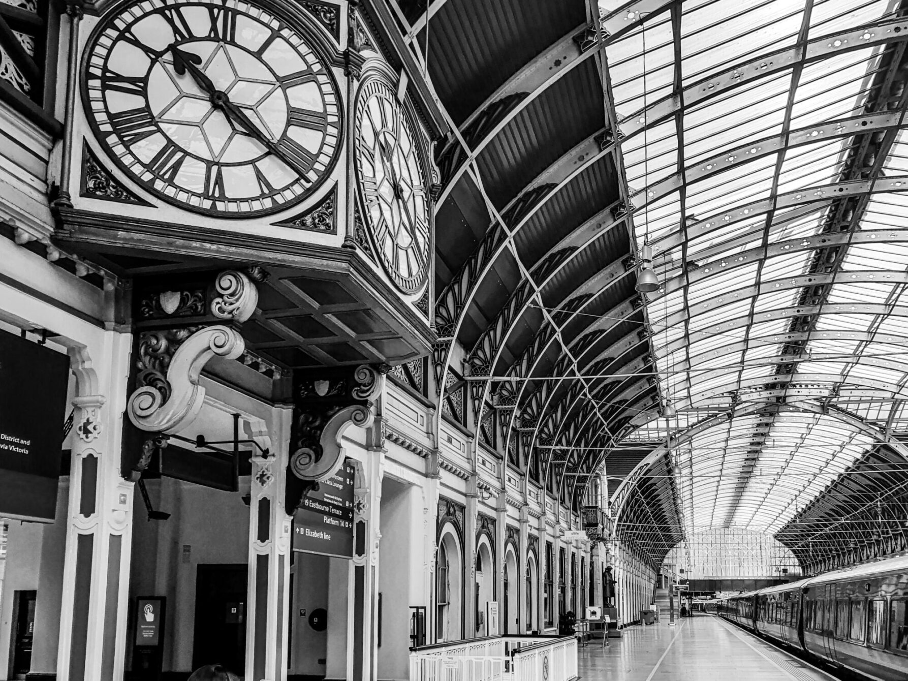 Black-and-white photograph looking up at a large ornate station clock beneath a curved glass-and-iron canopy roof, with platform signage and a train visible in the distance.