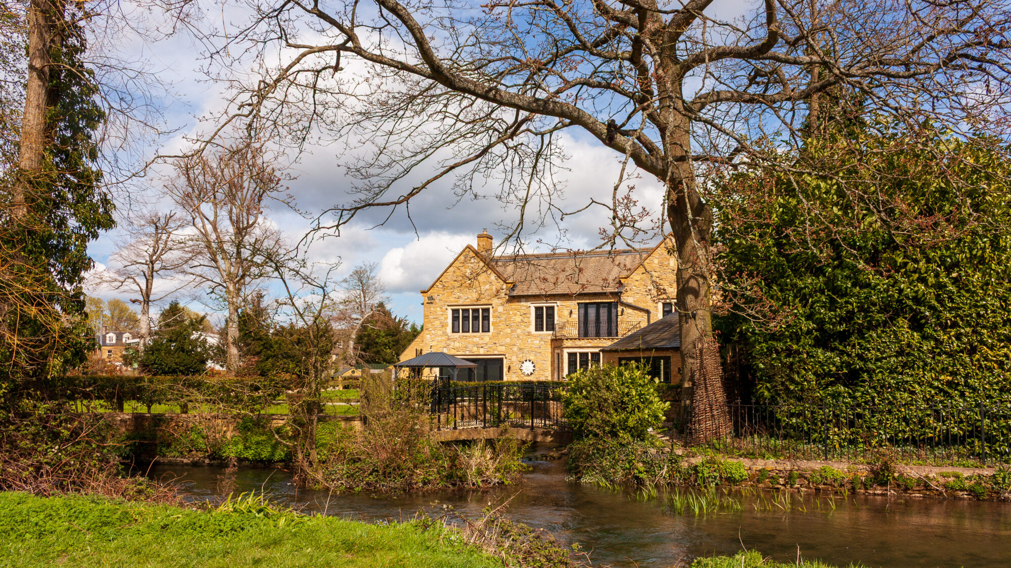 A honey-coloured stone Cotswold house with gabled roofs, seen across a small stream spanned by a footbridge, framed by bare tree branches and a blue sky with scattered clouds.