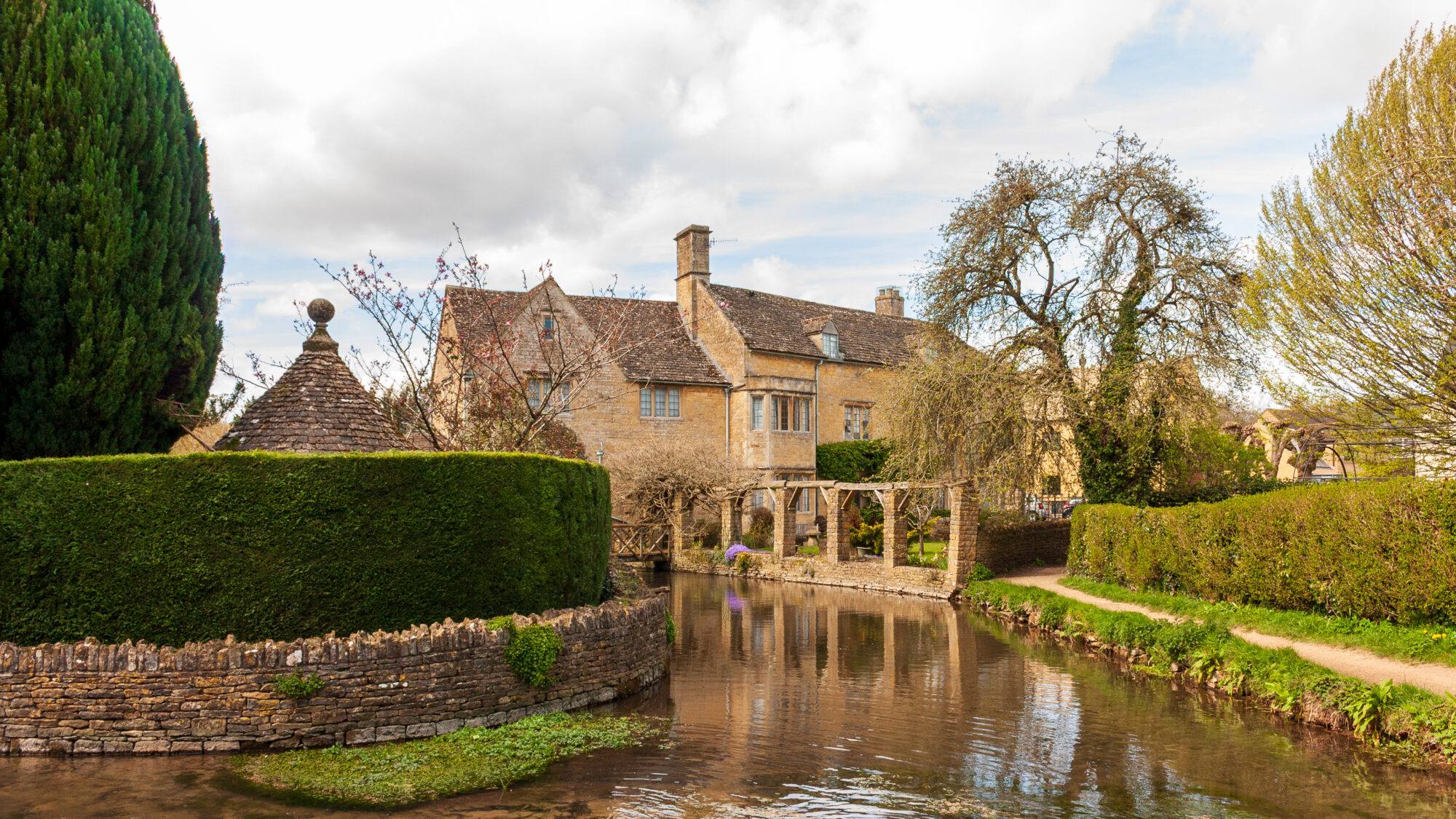 A traditional Cotswold stone manor house with tall chimneys and a stone pergola, reflected in a slow-moving stream bordered by neatly trimmed hedges and a dry-stone wall.