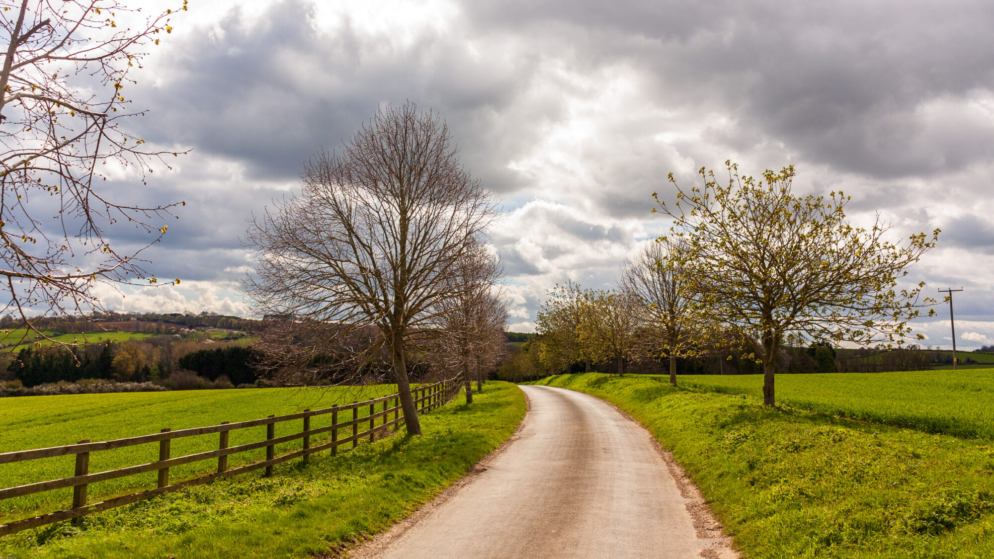 A quiet country lane lined with young trees and a wooden post-and-rail fence, running through green fields under a dramatic sky filled with grey and white clouds.