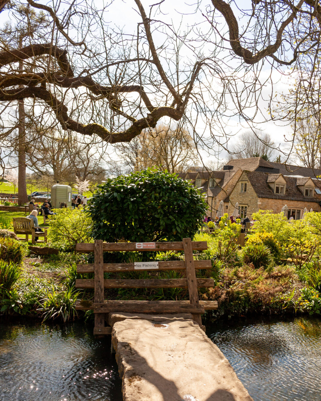 A rustic wooden footbridge with "No entry" and "No fishing" signs crossing a stream, with a Cotswold stone building, garden seating and visitors in the background.