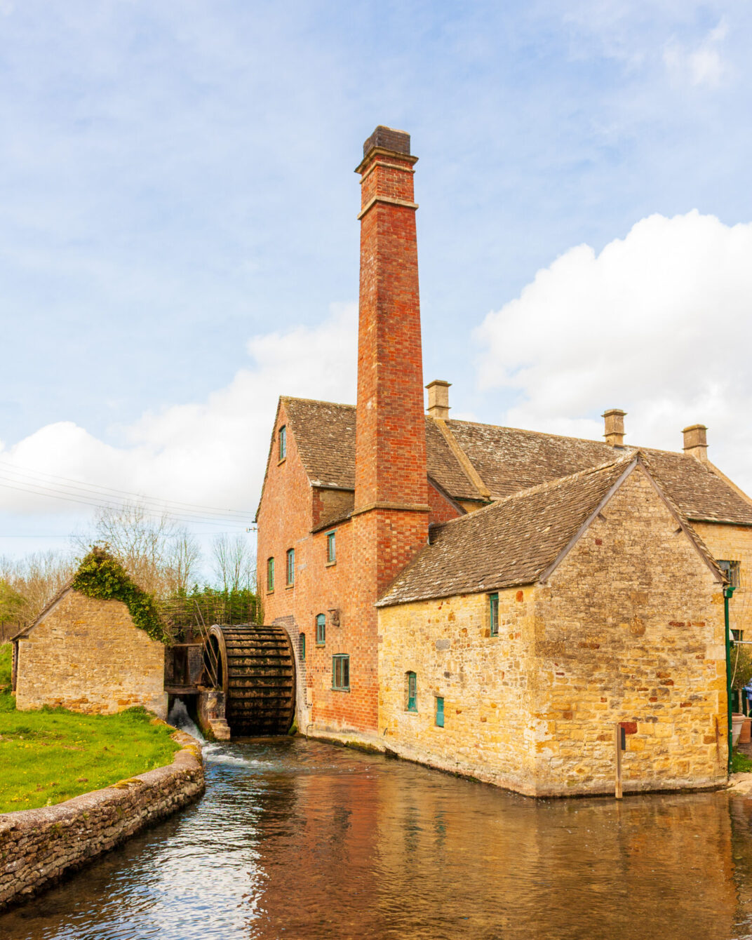 An old stone and brick watermill with a tall chimney and working waterwheel, set beside a stream, under a blue sky with soft clouds.