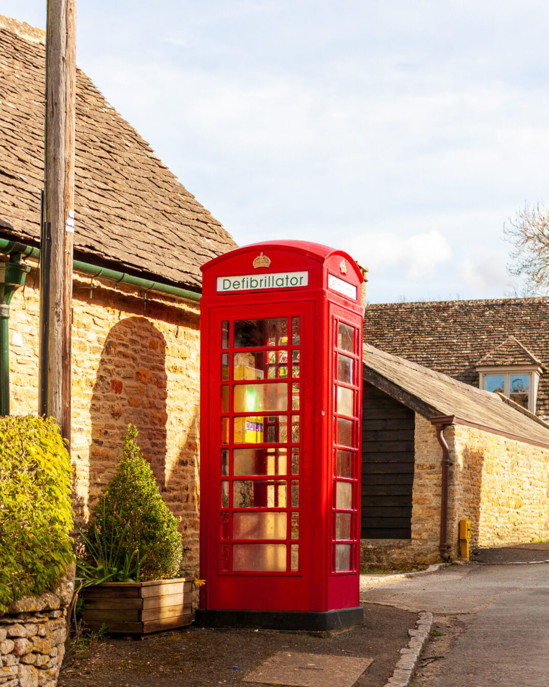 A traditional red British telephone box, repurposed as a defibrillator station, standing beside a stone cottage with a stone-tiled roof.