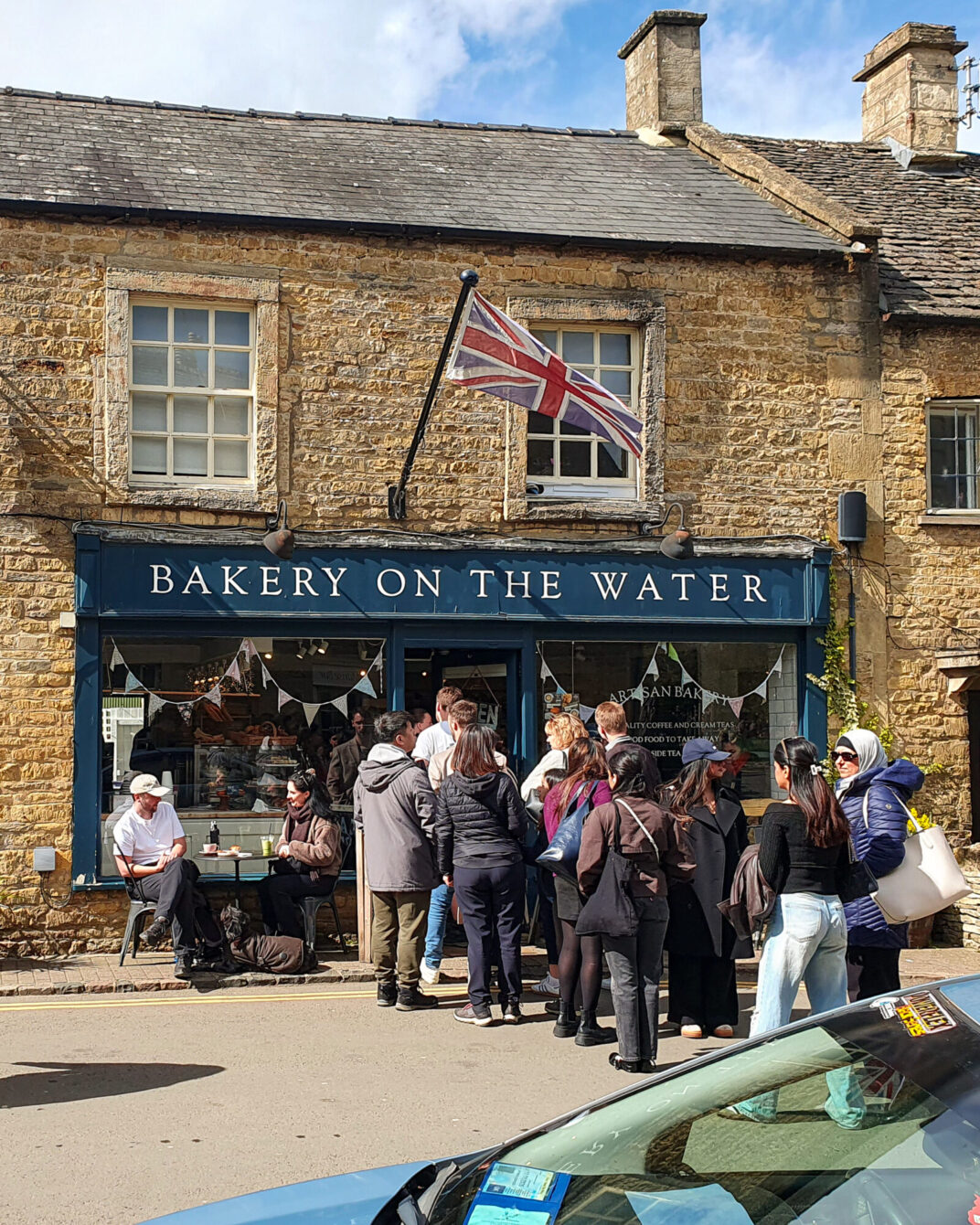 A queue of visitors outside "Bakery on the Water," a stone-fronted bakery with a Union Jack flag flying above the shop sign, under a partly cloudy sky.