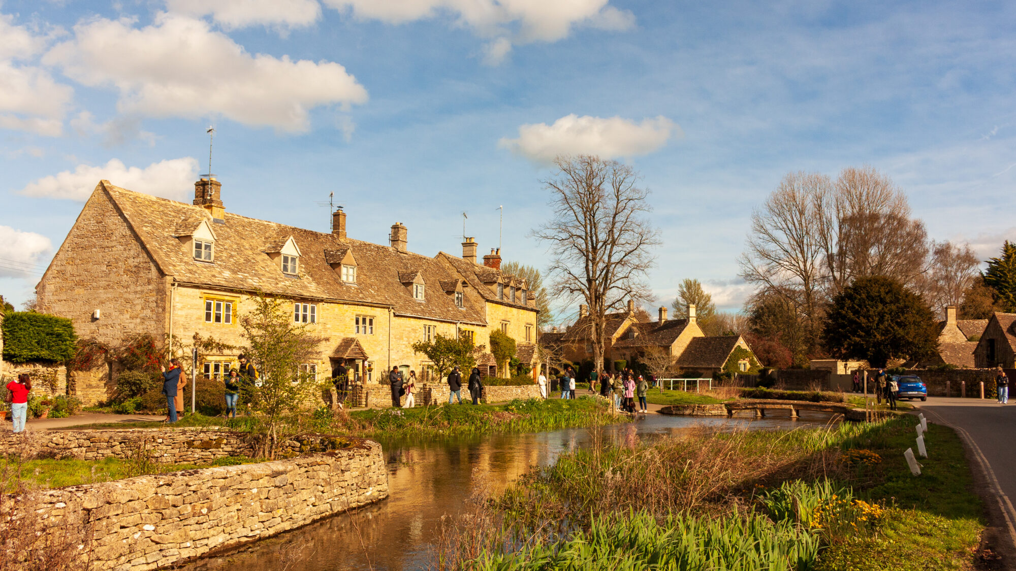 A row of golden Cotswold stone cottages with dormer windows, lit by warm evening sunlight, beside a stream with a small footbridge and visitors walking along the bank.