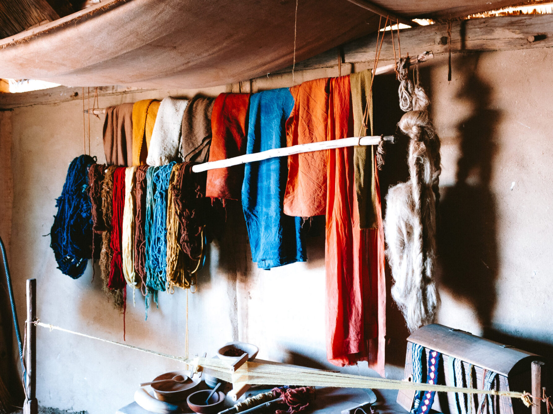 Skeins of hand-dyed wool and lengths of dyed cloth in shades of blue, red, orange and ochre hanging inside a Viking-age house, with a small hand loom and dye bowls in the foreground.