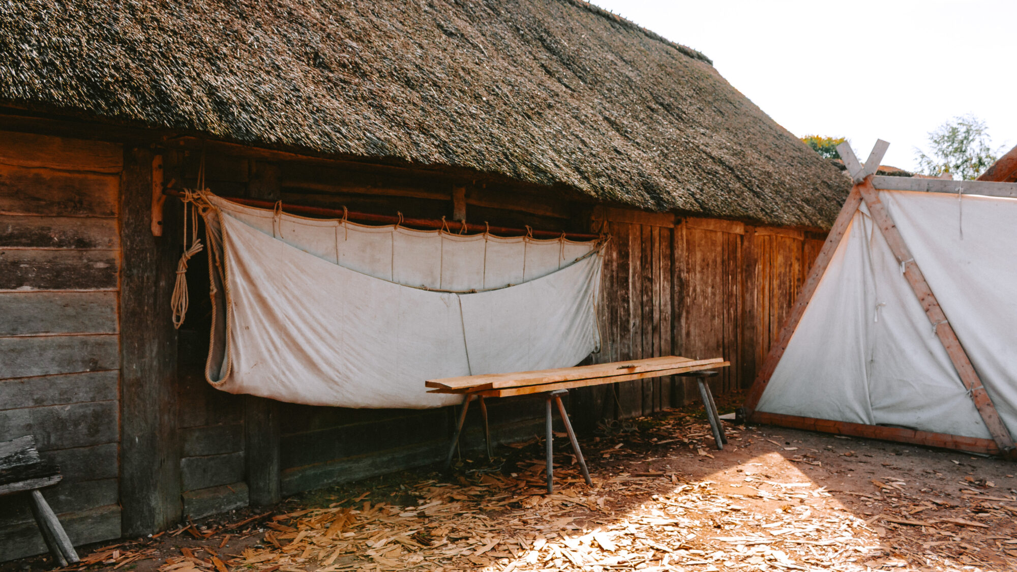 A wooden workbench and canvas awning outside a timber-and-thatch workshop building, with wood shavings scattered on the ground and a small canvas tent alongside.
