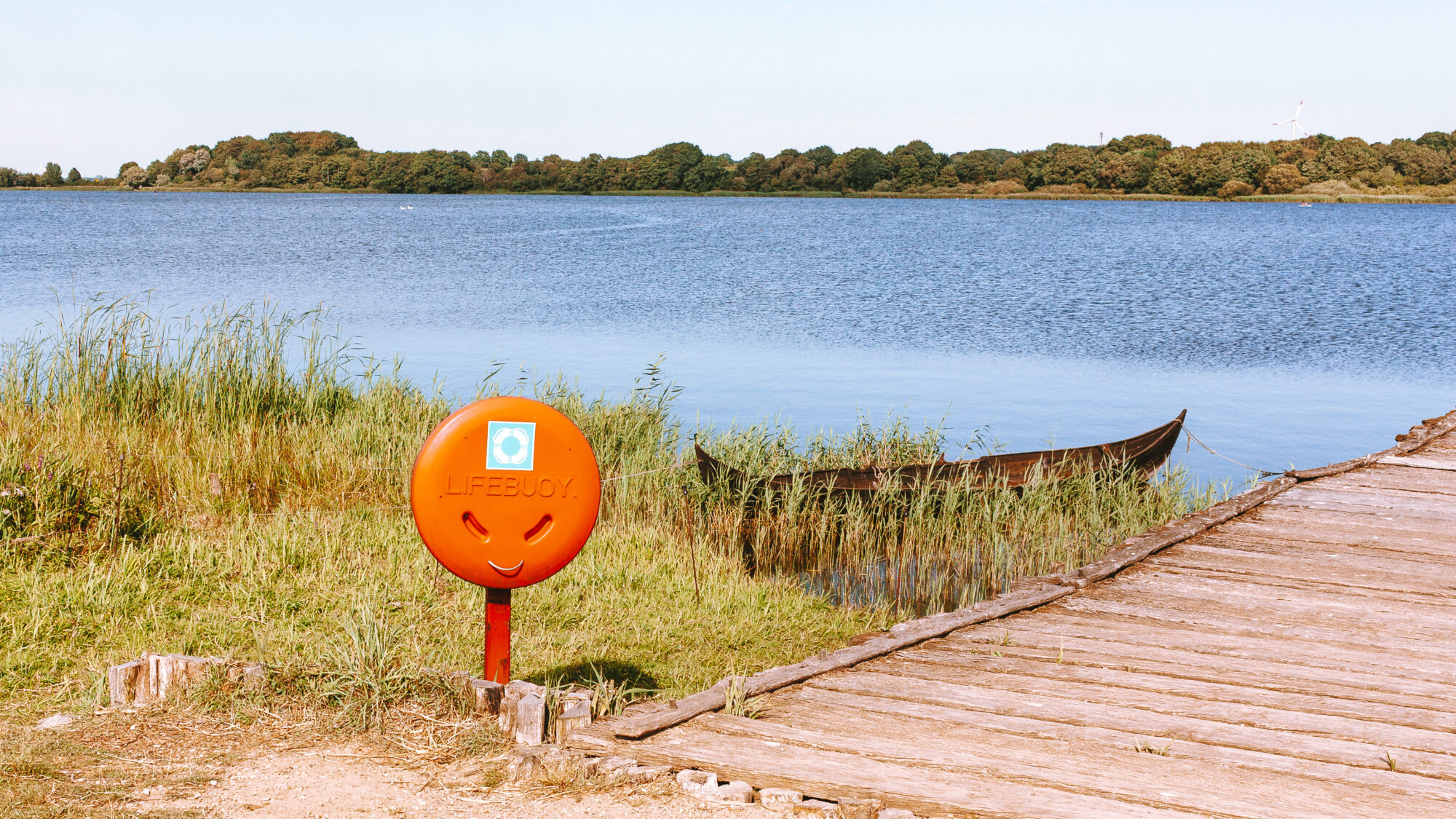A bright orange lifebuoy holder shaped like a smiling face standing beside a wooden jetty, with an old wooden boat resting in the reeds and a calm lake stretching to a wooded shoreline beyond.